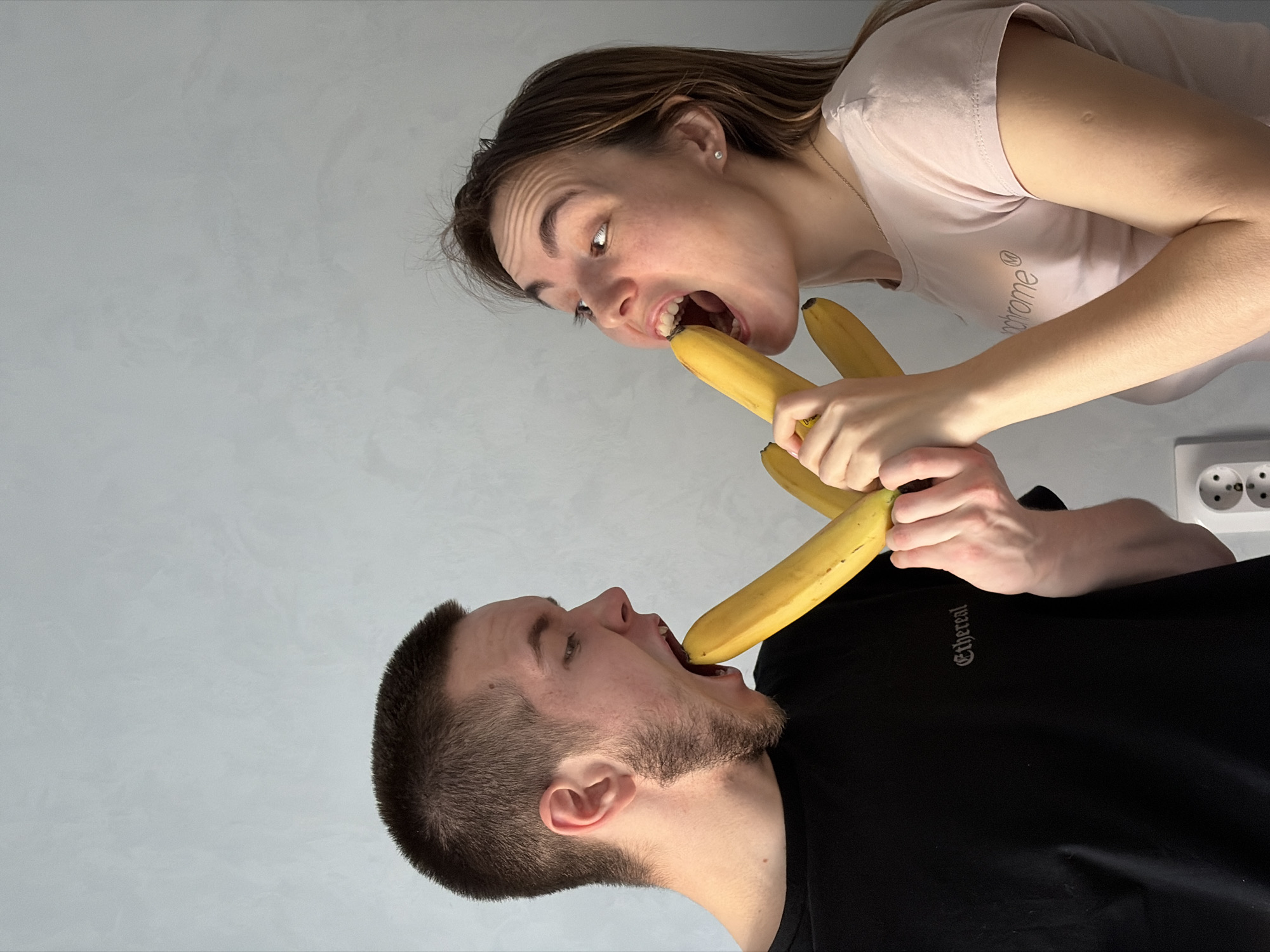 Two individuals, a male and a female, are interacting with bananas. The male is wearing a black shirt with the word "Ethereal" printed on it and has a banana partially in his mouth. The female, in a light pink shirt, is holding a bunch of bananas and also has one partially in her mouth. The location appears to be indoors with a white wall and electrical outlets visible in the background. There is no direct indication that the location is in Sarapul, Russia.
