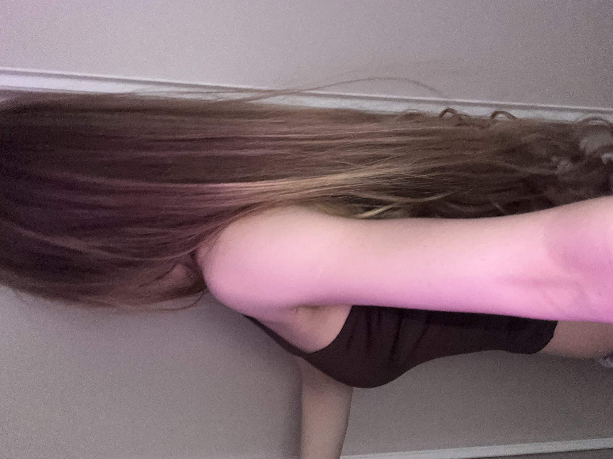 The image displays a close-up, rotated view of a person indoors. The subject is primarily defined by their long, multi-toned hair, which ranges from dark brown near the roots to lighter, brownish-blonde towards the ends, showing both straight and slightly wavy textures. The hair flows across the upper portion of the frame (as presented).
A prominent feature is the person's right arm, extended forward, suggesting they are holding a camera or phone to take the picture. This arm, along with parts of the shoulder and hair, is illuminated by a strong pink or magenta light source, giving it a distinct hue. The person is wearing a dark, form-fitting top, possibly a tank top or sports bra, which is visible on their upper torso. A small portion of what appears to be a knee or lower leg is visible at the bottom right.
The setting is an interior space, characterized by a plain, light-colored wall in the background, possibly off-white or light grey, with a subtle line indicating a seam or corner. The lighting appears artificial due to the pink glow. No specific activity beyond posing for or taking a photograph can be discerned. There is no visible text. The image's perspective is from a low angle, or the subject is oriented horizontally, with the image rotated 90 degrees clockwise from what would be a natural upright posture. Given the location context of Novoe Devyatkino, Russia, the scene depicts a casual indoor moment.
