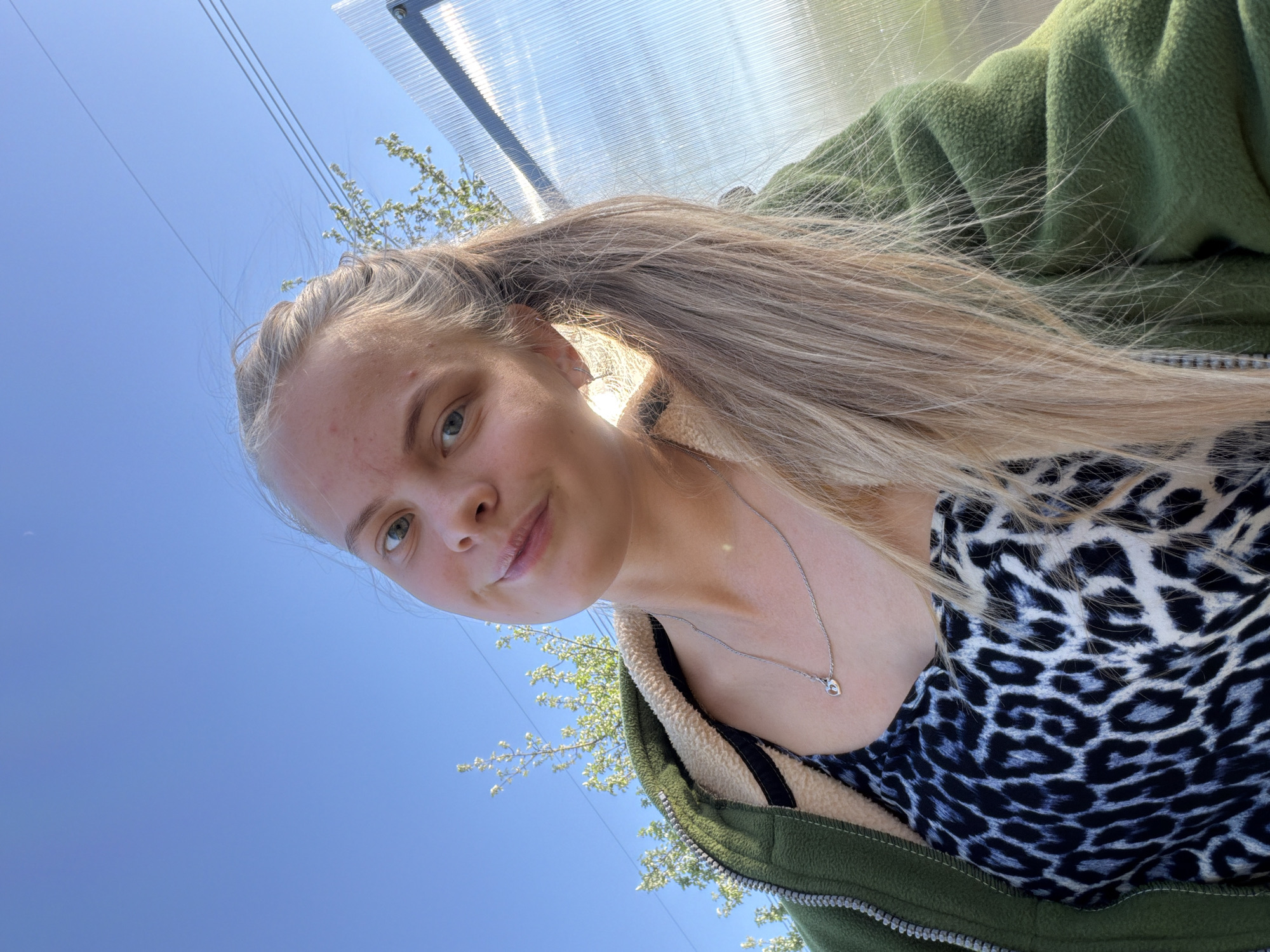 A fair-skinned woman with long, light brown hair in a high ponytail is facing the camera. She is wearing a black and white animal print top and a green fleece jacket. A silver necklace with a small pendant is visible around her neck. The background is a clear blue sky with tree branches visible in the bottom left corner. A translucent structure with visible frame elements is visible at the top. The photograph appears to have been taken from a low angle. There is no information to discern if the picture was taken in the city of Chaikovsky, Russia.