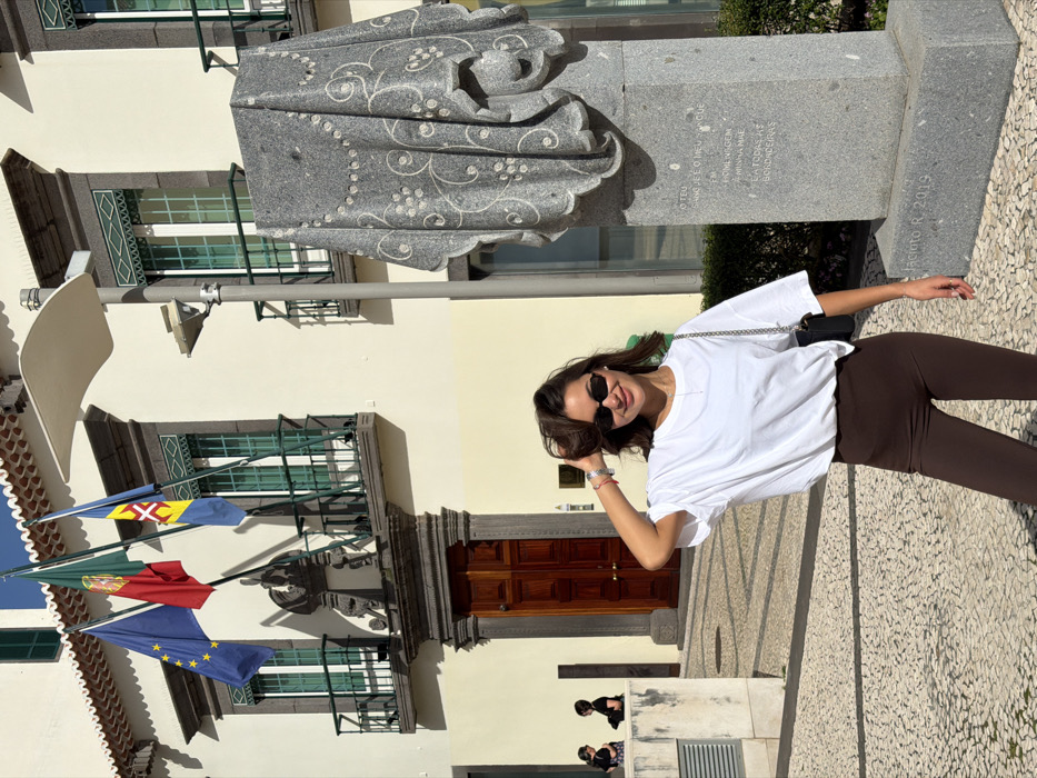 The image captures a vibrant, sunlit outdoor scene, likely in a town square in Madeira, Portugal. The main subject is a woman posing happily in front of a stone statue and a building adorned with flags.
The woman is positioned slightly to the right of the frame, smiling broadly at the camera. She has shoulder-length dark brown hair and is wearing black sunglasses, a loose-fitting white short-sleeved t-shirt, and dark brown or black leggings. A small black shoulder bag with a silver chain strap is visible across her body. Her left arm is bent, with her hand lightly touching her hair, conveying a casual pose. She also wears a watch on her left wrist.
Behind the woman and to her left stands a large, grey stone statue. The statue depicts a figure, possibly a woman, enveloped in an intricately carved drapery that features elaborate swirling and floral patterns. The base of the statue is rectangular and also made of grey stone, with text inscribed on its front and side. Visible text includes phrases in Portuguese such as "O TEU SANGUE É O MEU CÚ...", "FEMININA MÃE", "A MINHA MÃE", and "TODAS AS BORDADEIRAS" (meaning "All the Embroiderers"). Below this, parts of "DIAS DE..." and "2019" are discernible. On the lower right side of the statue's base, "JACINTO R. 2019" is clearly visible. Two blurry figures, possibly people sitting or resting, can be seen in the background near the base of the statue.
The background features a light-colored, cream or pale yellow building with multiple floors. It has dark green-framed windows and balconies with dark grey stone supports and green railings. At ground level, there is a large, ornate dark brown wooden door, suggesting an entrance to a significant building. Three flags are displayed prominently on poles attached to the building's facade: the top flag is that of Madeira (blue, yellow, blue with a red cross of Christ on a white shield), the middle flag is that of Portugal (green and red with the national coat of arms), and the bottom flag is the European Union flag (blue with a circle of 12 yellow stars).
The ground is paved with a distinctive pattern of light-colored, irregularly shaped stones, typical of Portuguese pavement (Calçada Portuguesa). The bright sunlight casts strong shadows, indicating a clear, sunny day, likely during the mid-morning or afternoon. The overall atmosphere is pleasant and relaxed.