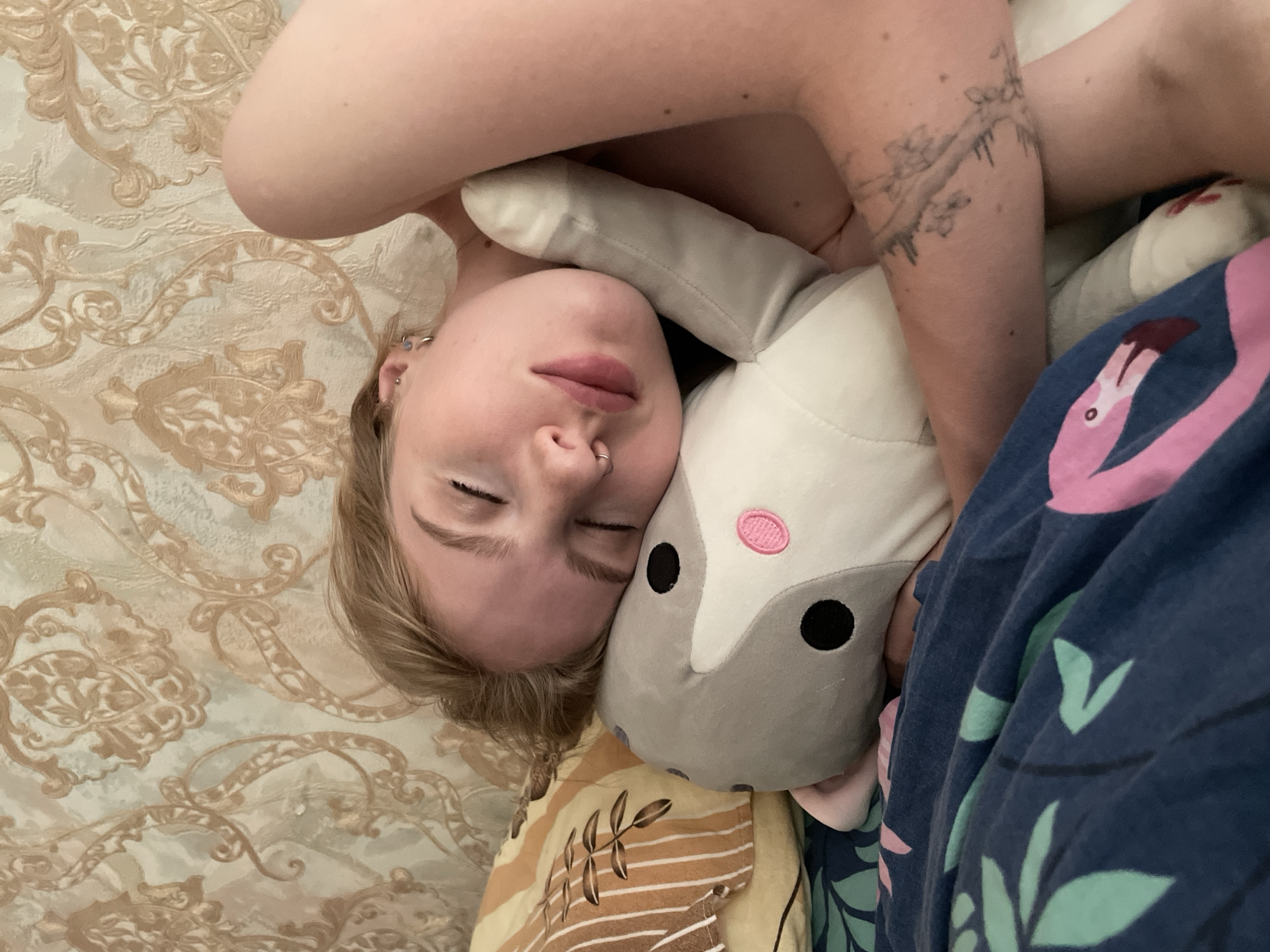A person is lying down, face towards the camera, embracing a gray and white plush toy. The person has fair skin, light blonde hair, closed eyes, a nose ring, and multiple ear piercings. A tattoo is visible on the person's left arm. The plush toy has black eyes and a pink nose. The background includes a patterned beige and brown surface, possibly a bedspread, and a blue patterned blanket with pink and green designs. There is no indication of the location being Izhevsk, Russia.