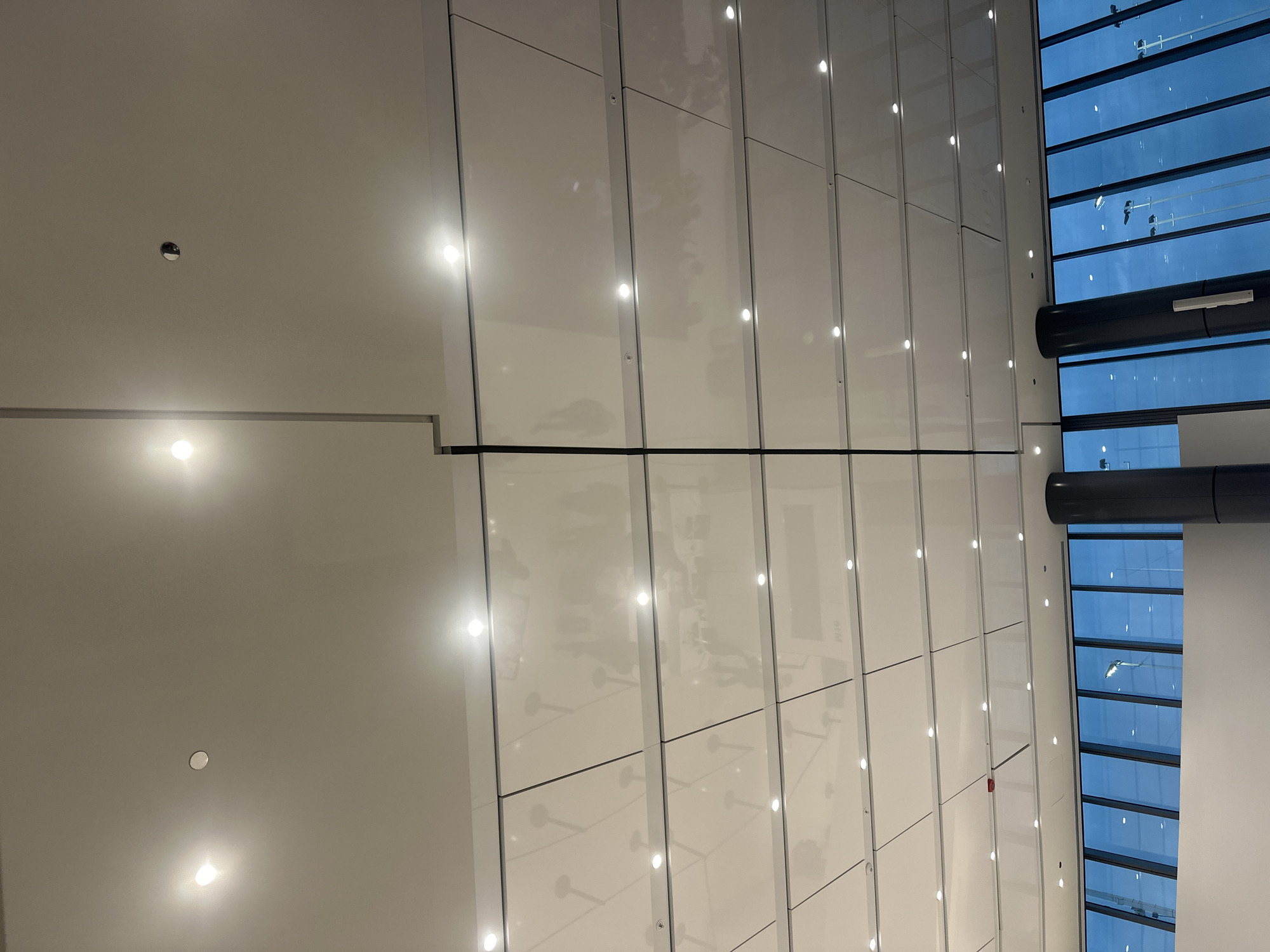 This image captures a modern interior with a sleek, white, modular ceiling featuring numerous recessed spotlights. The ceiling panels are arranged in a grid pattern, with some panels appearing slightly reflective, showing faint distortions of the overhead lighting. A prominent vertical dark line, possibly a seam or a structural element, bisects the ceiling from top to bottom.
Below the ceiling, a wide bank of floor-to-ceiling windows with a blue tint reveals an exterior scene at dusk or night. The windows are divided by vertical mullions. Through the glass, various streetlights or lighting poles are visible in the distance, suggesting an urban environment. Two large, dark gray cylindrical columns are present in the foreground, partially obscuring the view through the windows. The lower portion of the image shows a white wall.
While the image depicts an interior space with an external view, there are no people or specific identifiable objects within the scene that would definitively place it in Chicago, United States of America. The architecture and design are consistent with contemporary public spaces like airports, convention centers, or large retail establishments found in many major cities.
