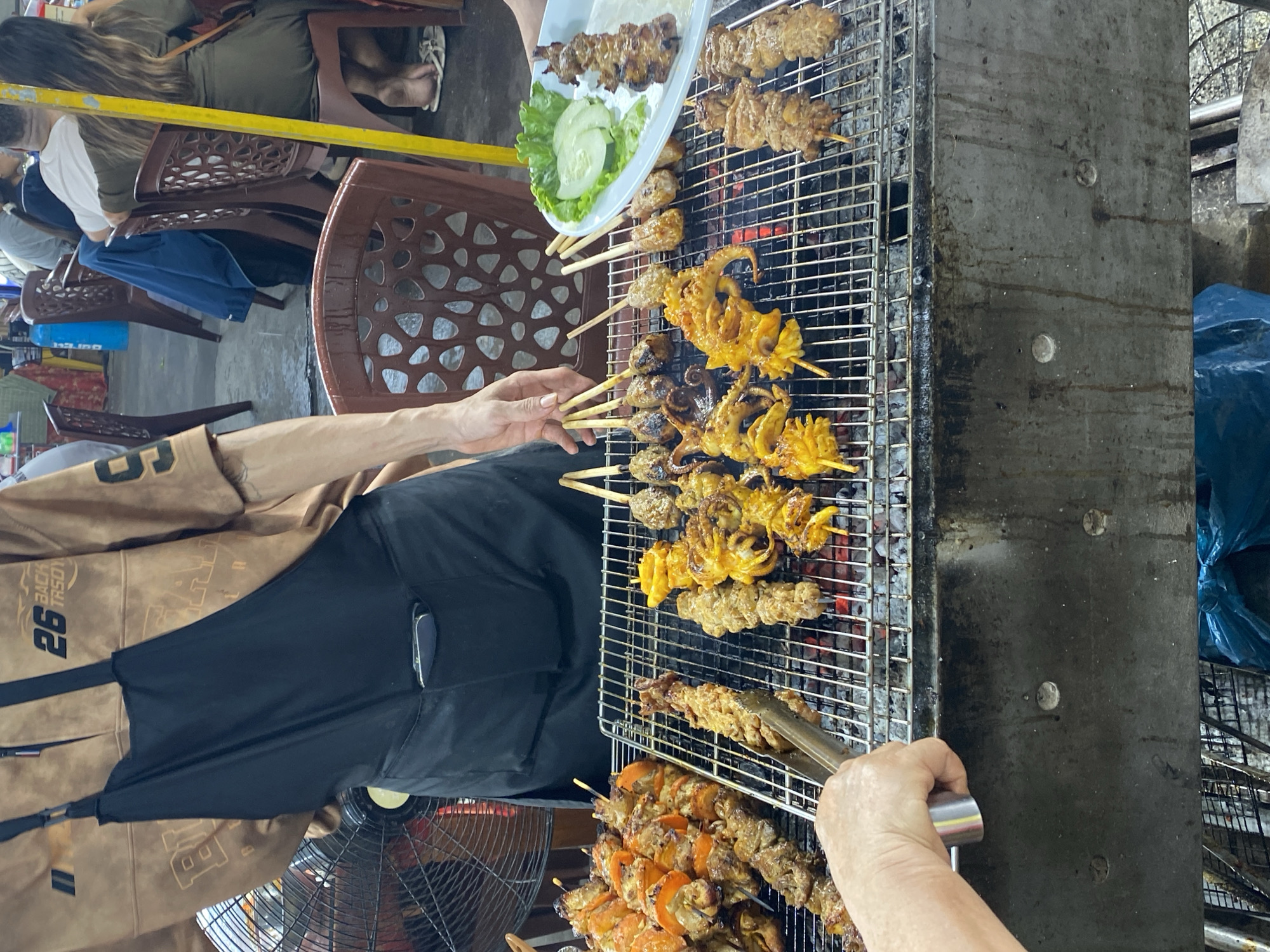 An outdoor street food stall in Da Nang, Vietnam, features a grill with various skewers of food cooking over hot coals. A person wearing an apron is tending to the grill, using tongs to adjust a skewer of meat. On the grill, there are skewers of what appear to be seafood, possibly squid, and meat. To the right, a white plate holds a skewer of grilled meat, along with lettuce and cucumber slices, suggesting it is ready to be served. In the background, other patrons are seated at tables, indicating a bustling atmosphere. The stall is set up on a paved area, typical of street-side food vendors.