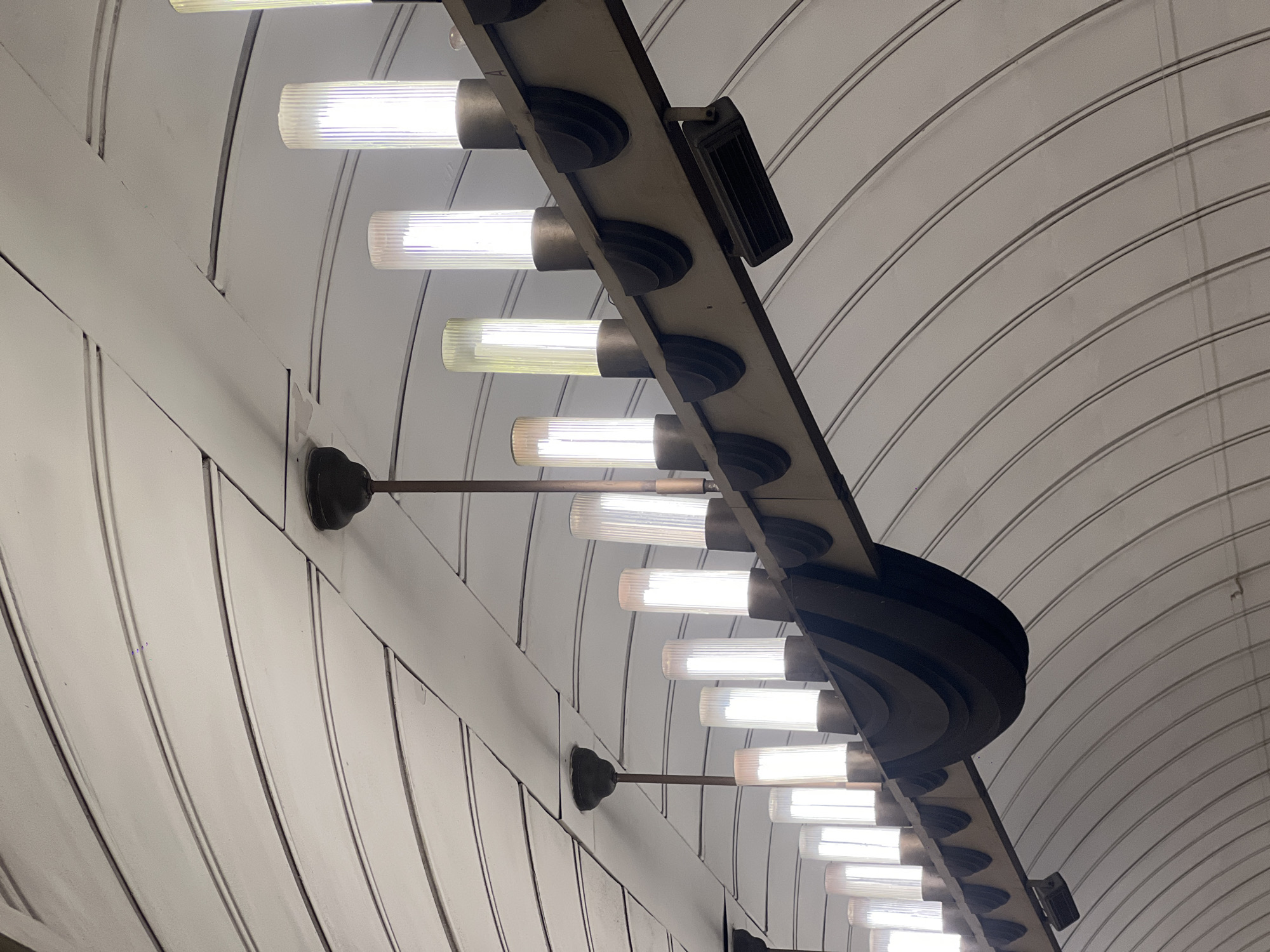 The image captures a view of the ceiling and lighting fixtures within a subway station. The location is likely in the city of London, United Kingdom, based on the architectural style of the station. The ceiling is constructed of arched, white panels with dark grey lines demarcating the segments. Running down the center of the frame is a series of light fixtures attached to a central support structure. Each fixture consists of a cylindrical, ridged glass or acrylic tube, emitting light. The support structure for the lights is a dark grey or black bar with decorative circular elements where each light is attached. At intervals along the support structure, there are additional decorative elements. The overall impression is one of a classic or Art Deco design often found in older subway systems.