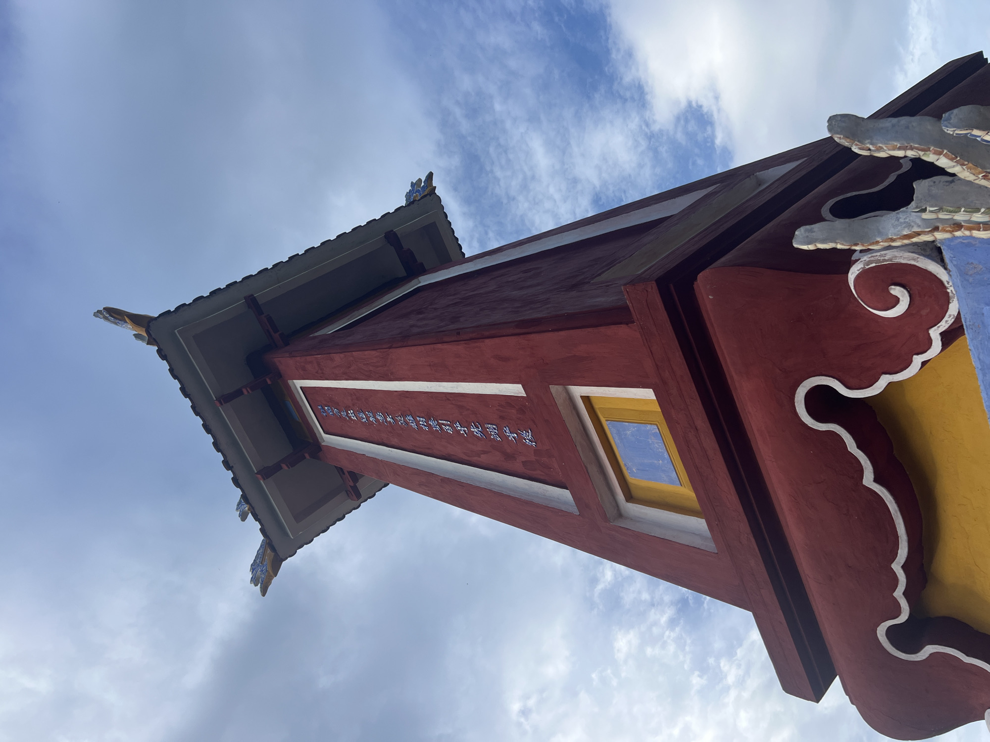 This is a low-angle, full-shot photograph of a traditional Vietnamese landmark in Hoi An, Vietnam. The landmark is a red, multi-tiered tower with a dark tiled roof and decorative elements. The tower is adorned with white and blue script on its upper section. A small, blue-and-yellow window is visible lower down. The base of the tower features ornate yellow and blue detailing. The sky above is blue with scattered white clouds. The photograph was taken from the ground, looking upwards, emphasizing the height and architectural details of the structure.