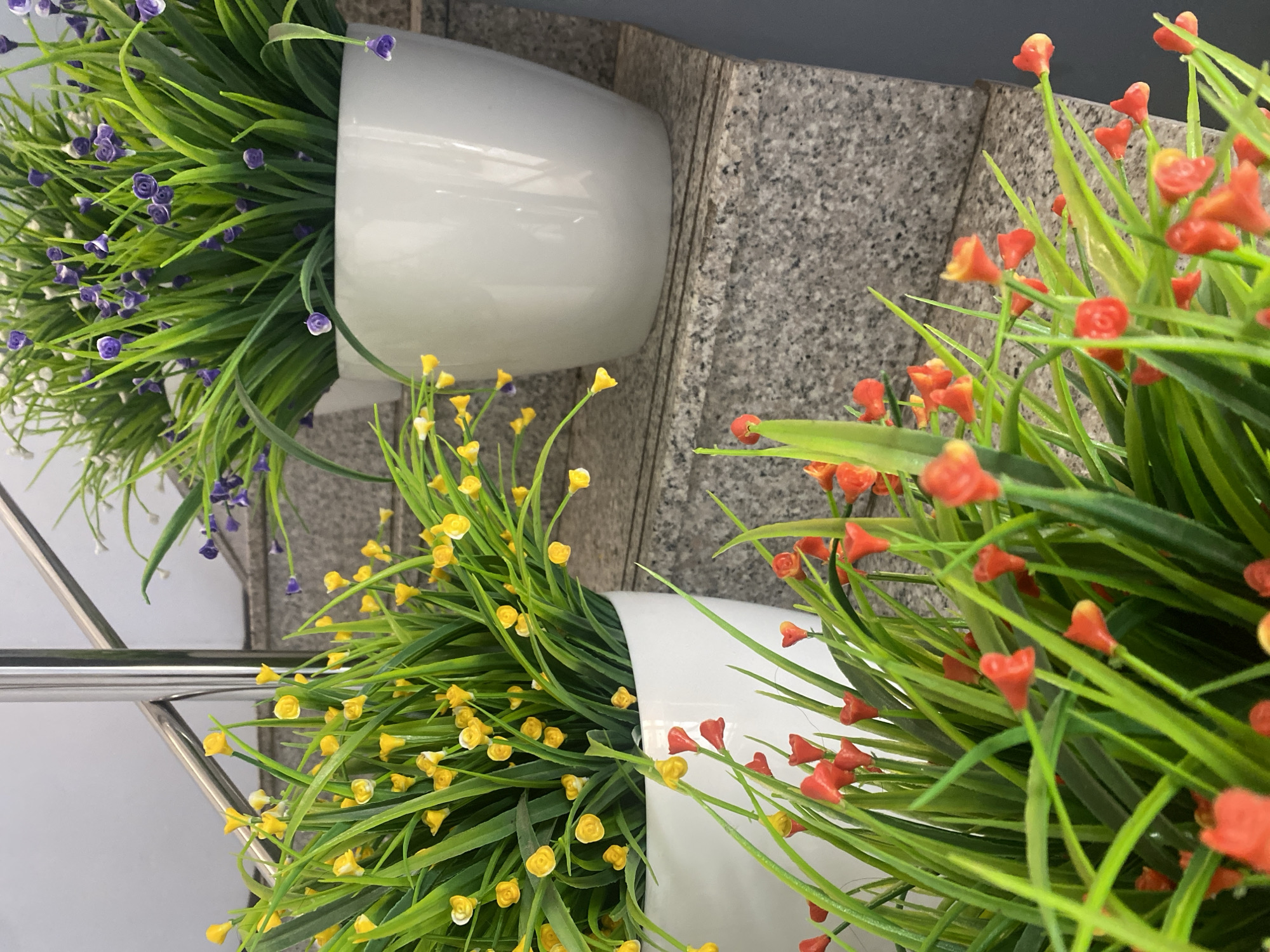 The image displays three potted artificial plants on a set of granite stairs. The plant in the foreground features green foliage with small red and orange flower-like accents. Behind this, to the left, is a pot with green foliage and small yellow flower-like accents. To the right and slightly further back is a pot of green foliage with small purple flower-like accents. A silver-toned handrail runs diagonally across the upper left portion of the image. The background consists of a light-colored wall. The scene is located on outdoor stairs in Astana, Kazakhstan.