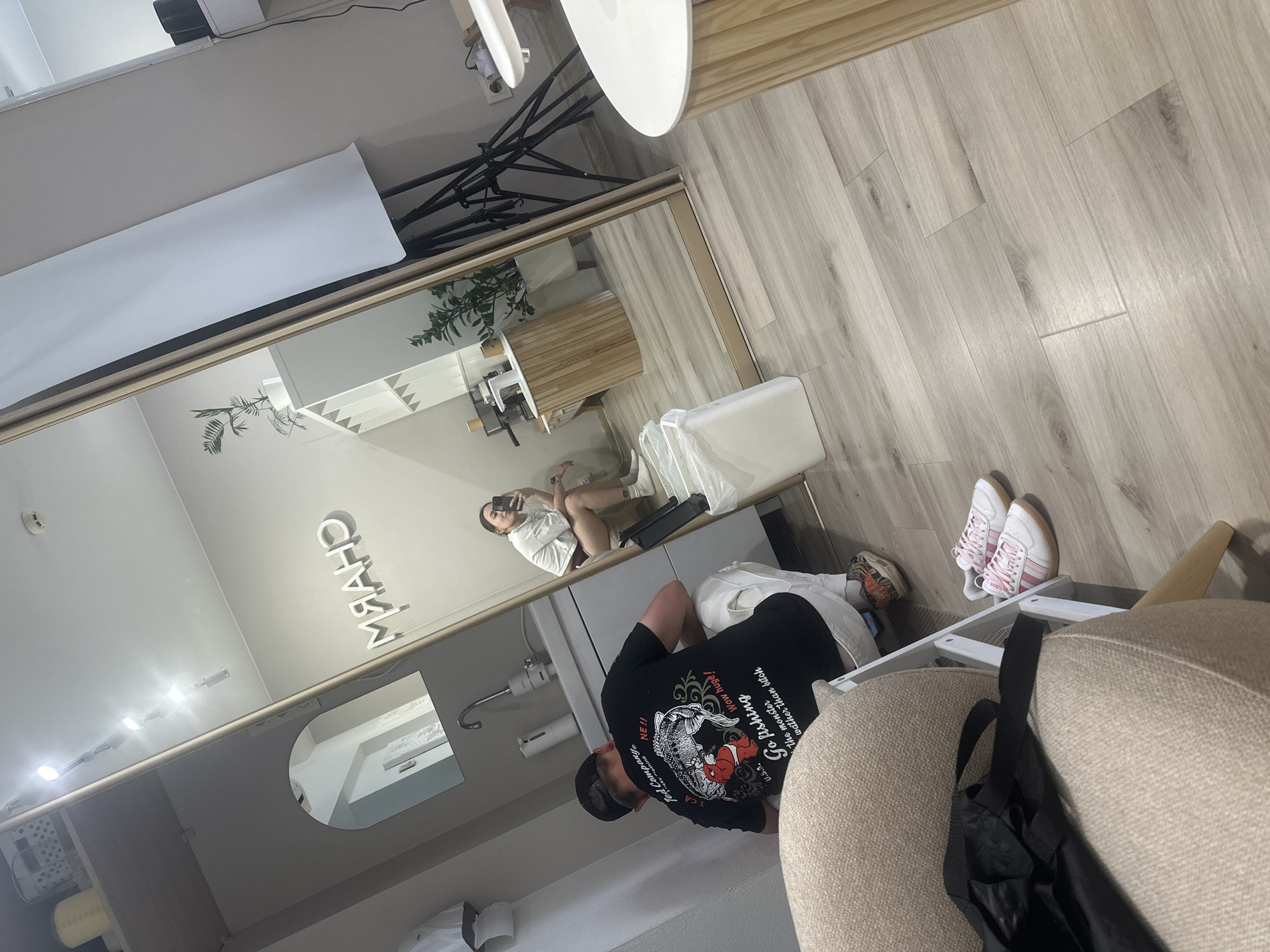 Here's a detailed description of the image:
**Setting:** The image appears to be taken inside a modern, brightly lit commercial space, likely a beauty salon or barbershop.
**People:**
* One person, a male, is the main subject. He's wearing a black T-shirt with graphic print, shorts, and a cap. He's seated or kneeling near a counter, possibly interacting with the space.
* The reflection in a large mirror reveals another person, a female, taking a selfie with her phone.
**Objects:**
* Large mirror spanning the length of a wall.
* A salon counter with a sink.
* Salon lighting fixtures.
* A chair with a black bag hanging on it
* Two pairs of shoes
**Actions & Interactions:**
* The male subject is looking or interacting with something out of frame or within the shop space.
* The female subject is taking a selfie reflected in the mirror.
**Environment:** The space is clean, well-lit with white walls, and has light-colored wooden flooring. The mirror provides an illusion of depth and reveals the interior design and arrangement of the salon. The space appears to be organized for commercial purposes.
The image does not include information related to a specific location or the city of Perm, Russia. The provided description focuses solely on what is visibly present in the image.