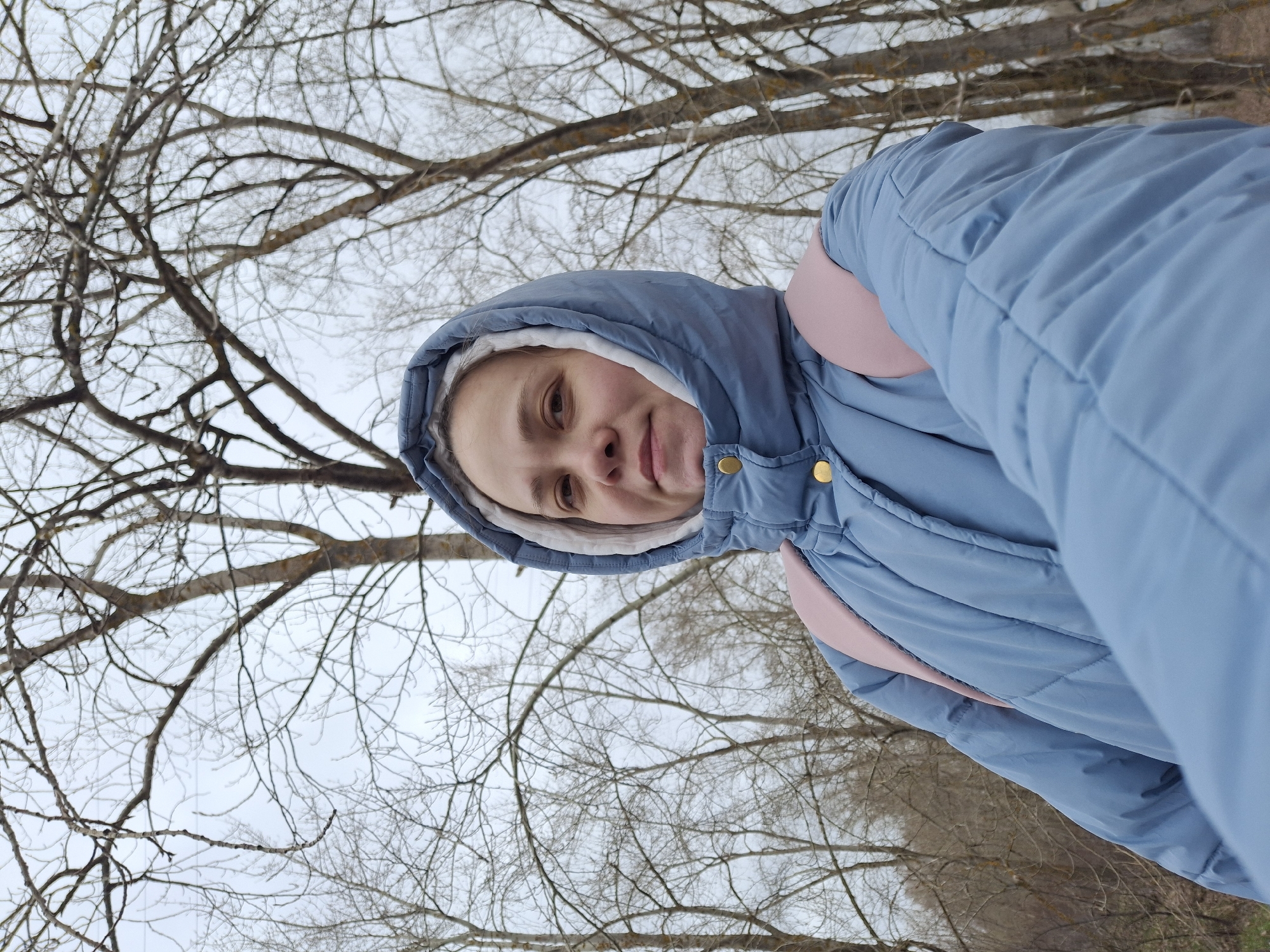 The image captures a selfie taken outdoors, featuring a young woman or teenager. The photo is rotated, showing the subject's head and upper torso on the right side of the frame, looking towards the left. She is wearing a light blue puffer jacket with the hood up, which has a white or light cream lining. Brass-colored buttons or snaps are visible on the jacket's front. A pink strap, likely from a backpack, is visible over her right shoulder. The subject has light skin and displays a subtle, pleasant expression or a small smile.
The background is dominated by a dense network of bare tree branches against an overcast, light grey sky. The absence of leaves on the trees indicates that the photograph was taken during late autumn, winter, or early spring. This setting suggests a wooded area, park, or forest, consistent with the location context of Chaikovsky, Russia, during colder months. The overcast sky implies a cloudy day, and the puffer jacket suggests cold weather conditions. No visible text is present in the image.