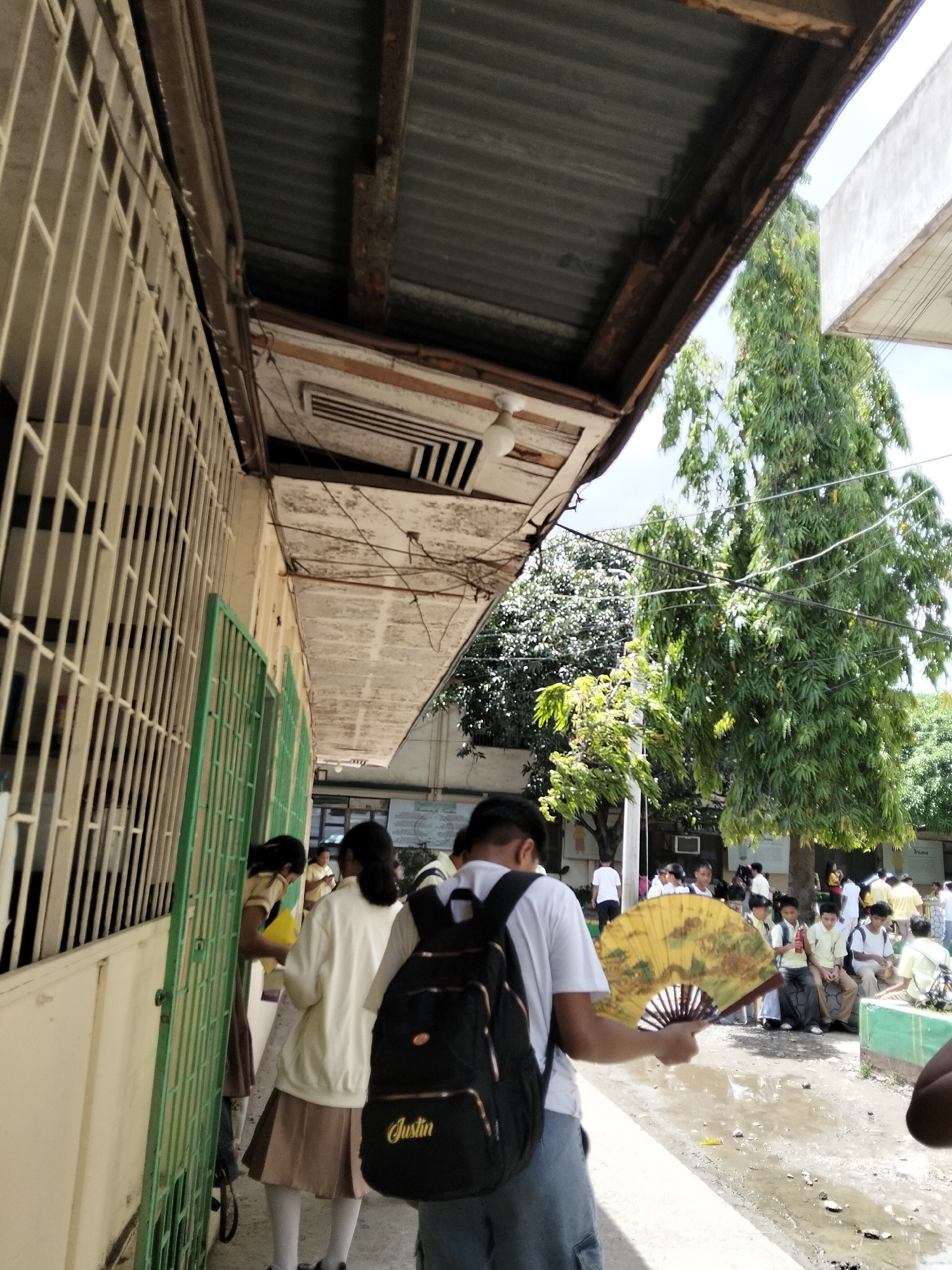 In an outdoor setting that appears to be a school in Davao City, Philippines, a group of students are visible. The architecture suggests an older building with a corrugated metal roof and a long, covered walkway with metal grates and green-barred windows. The ground is unpaved and wet, with puddles indicating recent rain.
In the foreground, a young male student, seen from behind, is wearing a white t-shirt, denim shorts, and a black backpack with the name "Justin" in cursive script. He is holding a large, open yellow fan decorated with a landscape scene.
Further down the walkway, several female students in school uniforms are seen. One student in a white jacket and a brown skirt is holding a yellow document. Another student in a similar uniform is partially visible.
In the background, more students are gathered, some sitting and others standing. A tall tree with dense foliage is prominent on the right side of the image. The overall impression is a typical school day scene with students interacting and moving about the campus.