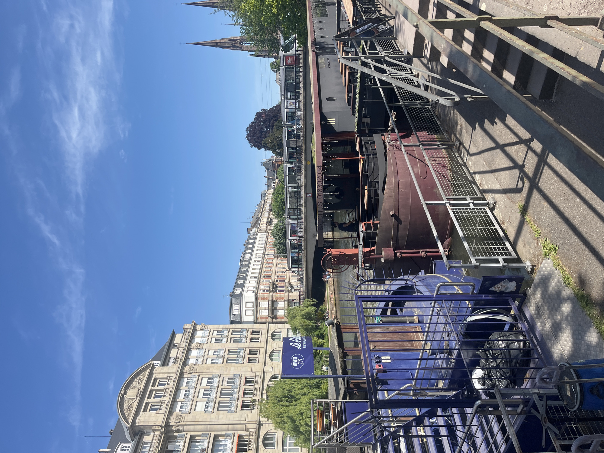 This media file depicts a vibrant daytime scene along a canal or river in Strasbourg, France. The image, if correctly oriented, shows a waterway running horizontally through the middle, flanked by buildings and infrastructure.
In the foreground, a blue structure, possibly a tour boat or part of a dock, features metal railings and what appears to be a black trash bag and other items inside. A dark red/brown barge is partially visible behind it. To the right, concrete steps and metal railings cast strong shadows, indicating bright sunlight.
The midground features the canal itself, where several boats are present. A long, dark barge with "ILLUSTRO" written on its side is prominent. Another boat, possibly a passenger vessel, is visible further down the canal. People can be seen on some of the boats and along the far bank. Lush green trees line the water's edge.
The background is dominated by stately, historic-looking buildings with multiple stories and ornate architectural details. One building exhibits a distinctive rounded facade. A blue sign clearly displays "Le Nôtre," "BAR BRASSERIE," and "OUVERT 7J/7," indicating a business. Rising above the rooftops, two distinct gothic-style church spires, characteristic of Strasbourg, punctuate the horizon.
The sky is a clear, bright blue with scattered, wispy white clouds, suggesting fine weather. The strong sunlight and clear skies indicate it is likely midday or early afternoon. Another piece of text visible on a boat or barge reads "EGUIS 018302034." The scene captures a lively urban environment, combining historic architecture with contemporary canal activity.