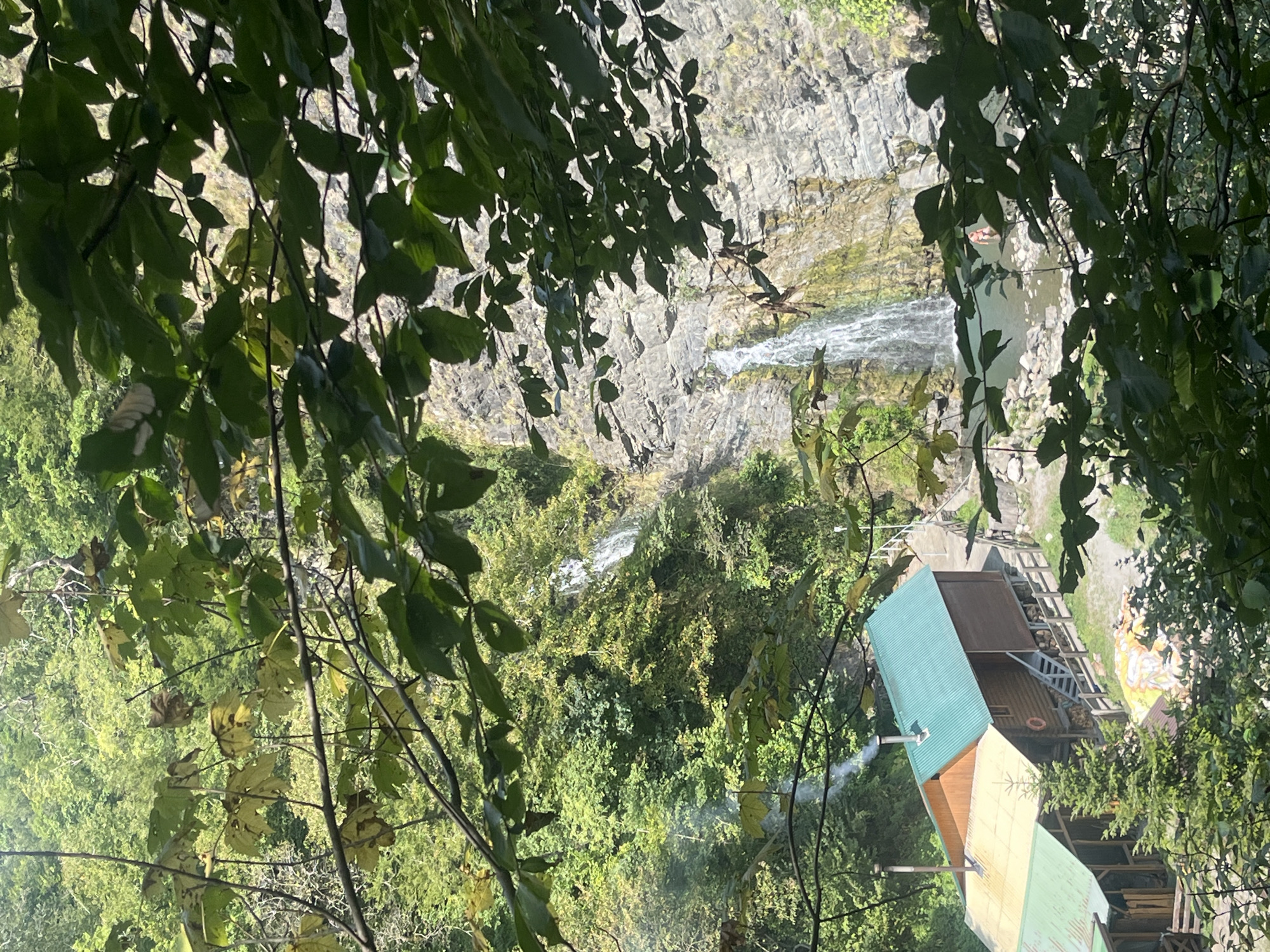 This image depicts a natural landscape featuring a waterfall cascading down a rocky cliff face. In the foreground, lush green foliage partially obscures the view, suggesting a dense forest environment. At the base of the waterfall, a body of water is visible, with a person swimming near the waterfall. To the left of the waterfall, nestled within the trees, are several wooden structures with green and yellow roofs. One of these structures appears to be a rustic cabin or lodge, with a chimney emitting smoke. The overall scene is one of a secluded natural setting, possibly a recreational area or park, with elements of human habitation integrated into the wilderness. The location is identified as Khostinskiy, Russia.