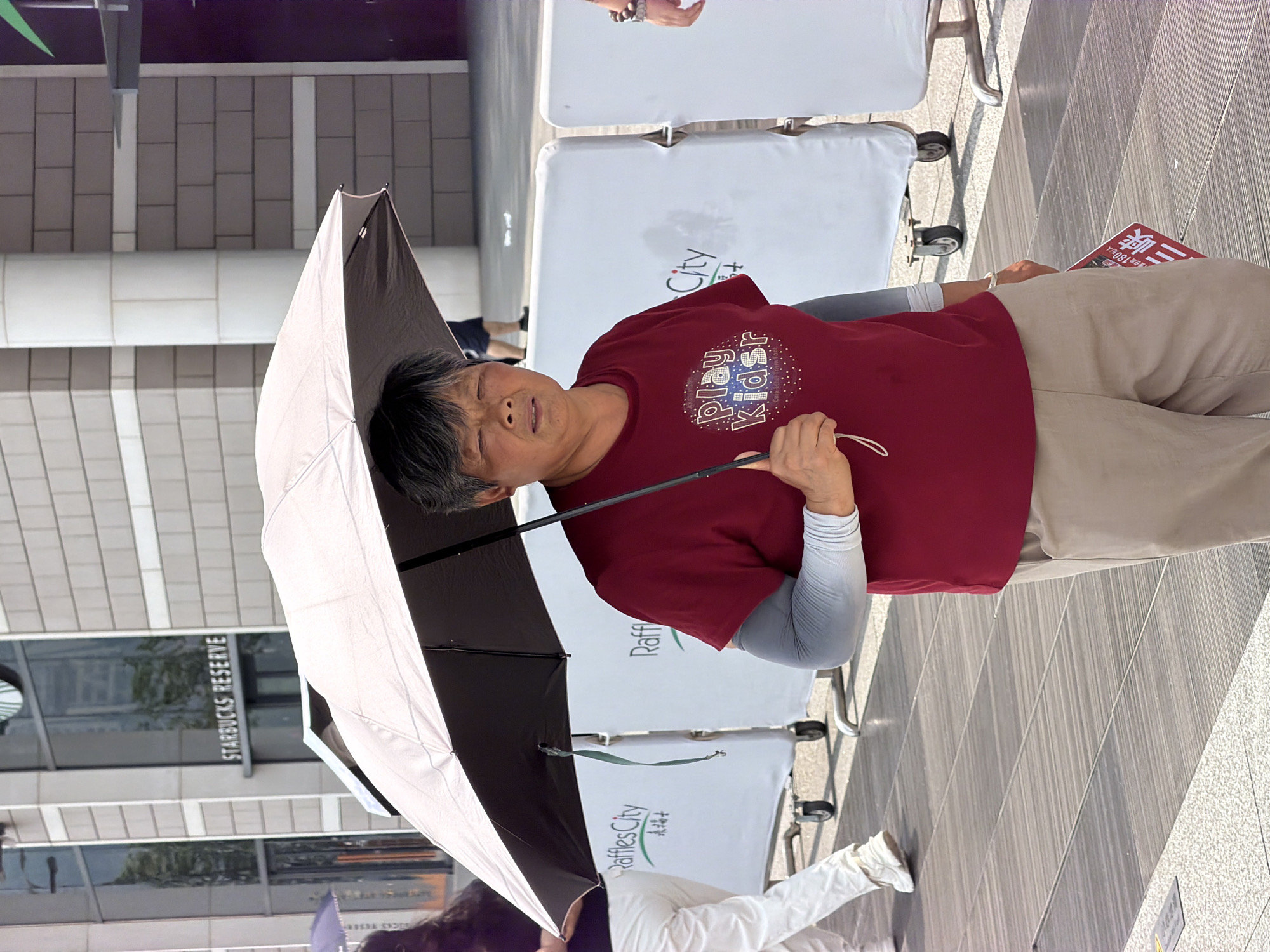 A middle-aged East Asian woman walks under a grey umbrella, holding the handle with her right hand. She is wearing a maroon t-shirt with a graphic that reads "play kidsr" in white letters, and a pair of light brown pants. She has on light grey arm sleeves. The woman's face is visible, looking forward with a neutral expression. Behind her, white barriers with "Raffles City" printed on them are visible. The background suggests an outdoor urban setting with a building featuring "Starbucks Reserve" signage. The ground is paved with grey tiles. The lighting indicates it is daytime.