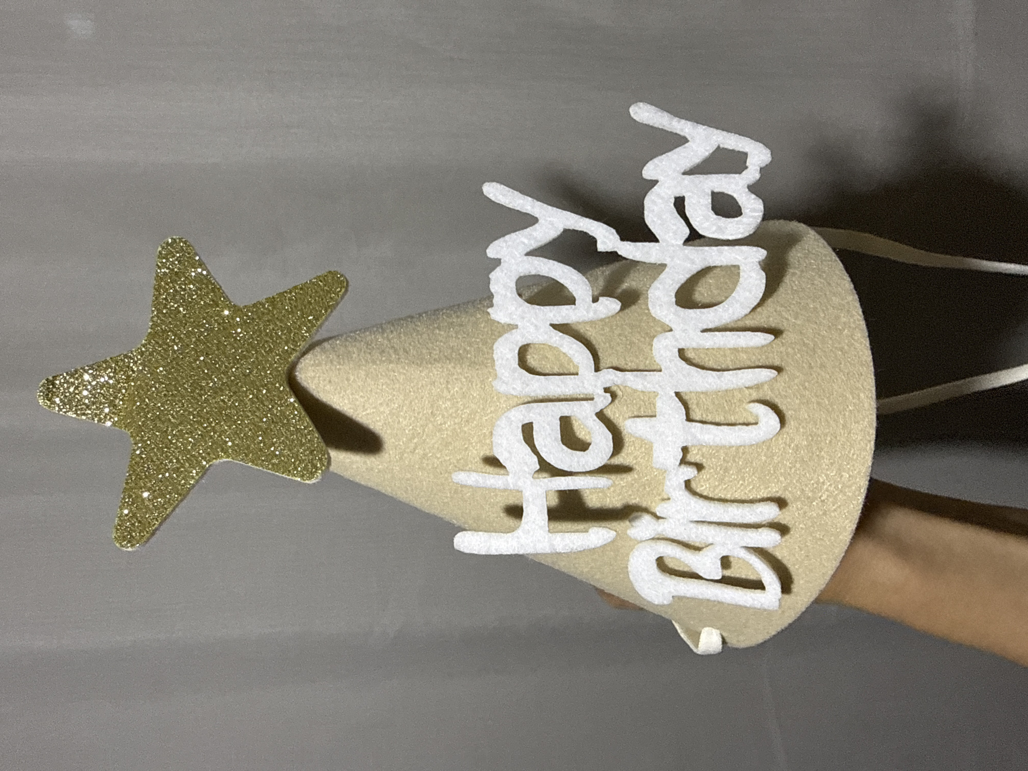 A person is holding a cream-colored birthday cone hat. The hat is decorated with a gold glitter star at the top and white felt lettering that spells "Happy Birthday." The hat has white elastic straps attached to it. The background is a plain gray fabric. The image appears to be taken indoors, with a single light source casting a shadow on the right side of the hat. There are no indications of the city of Nerudstal, Ukraine.