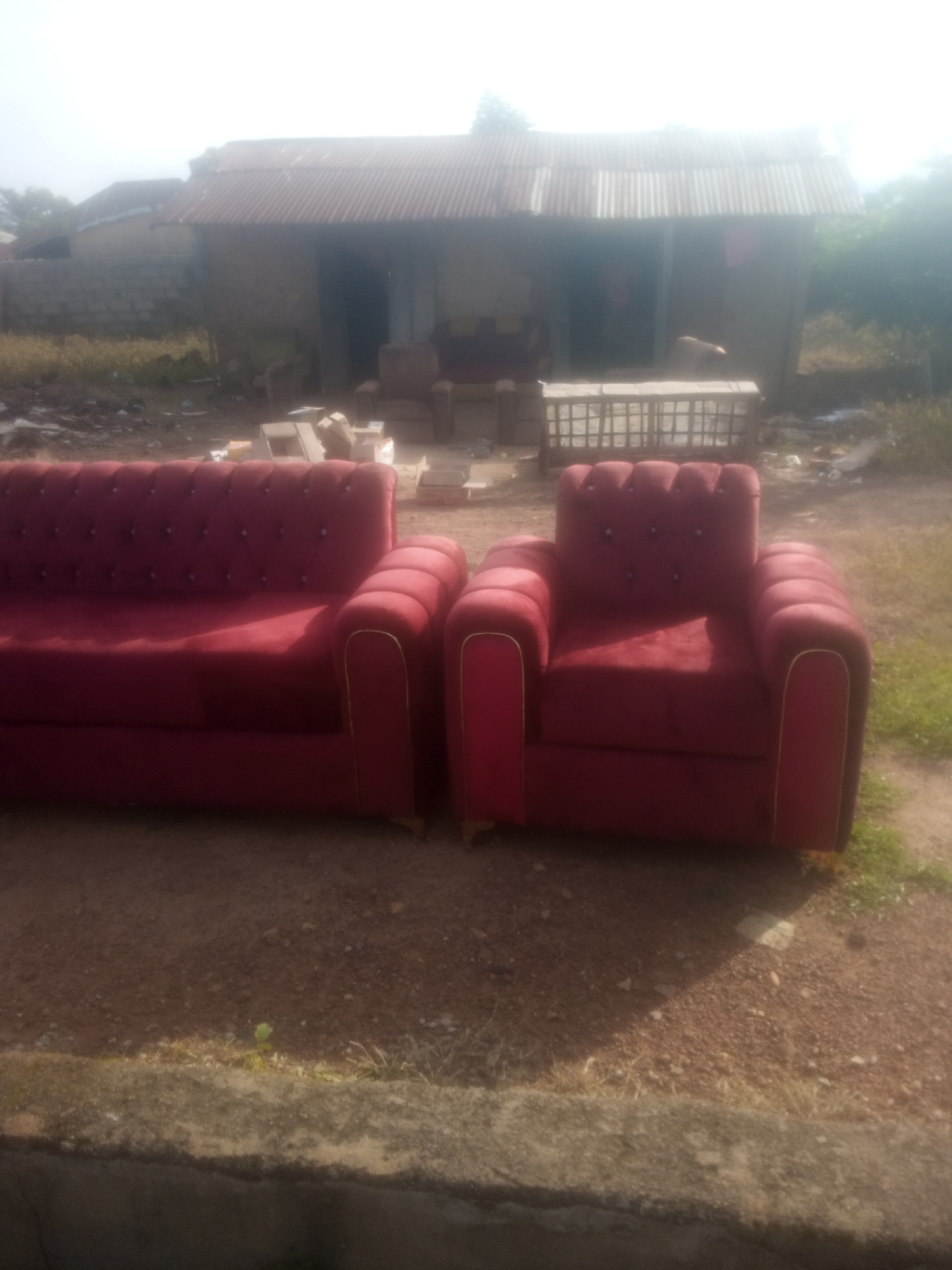 This image shows a red velvet sofa set placed outdoors in Serti, Nigeria. The set includes a two-seater sofa and a single armchair, both featuring tufted backs and rounded arms with gold trim. The furniture is positioned on a reddish-brown dirt ground, with dry grass and some scattered rocks.
In the background, a rustic building with a corrugated metal roof stands. The building appears to be a workshop or open-air structure, with a dark opening suggesting an entrance. Some piles of bricks and a wooden crate or shelf are visible near the building, indicating potential construction or storage activity. The overall scene suggests an informal setting, possibly a furniture maker's yard or a community space.
The lighting indicates it is daytime, with sunlight casting shadows on the ground and furniture. The weather appears to be clear, possibly sunny. The setting seems rural or semi-rural, given the natural ground cover and the simple construction of the background building. There is no visible text in the image. The image does not clearly depict any people or specific activities other than the placement of the furniture within its environment.
