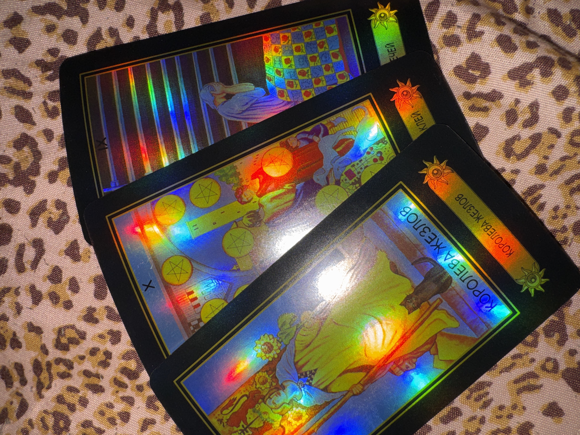 The image shows three holographic tarot cards laid out on a leopard-print fabric.
The top card, partially obscured, depicts a figure lying down, seemingly behind bars. The background is a checkered pattern.
The middle card displays a scene with multiple figures and several golden pentacles. One figure appears to be holding a cane or staff, and there is a white dog or similar animal.
The bottom card features a figure in yellow and blue robes, holding a staff adorned with sunflowers.
The text visible on the cards is in Cyrillic. On the top card, "ЕЕЙ" is partially visible. On the middle card, "КЛЕЙ" is visible. On the bottom card, "КОРОЛЕВА ЖЕЗЛОВ" is clearly visible, which translates to "Queen of Wands."
The holographic effect causes the cards to display a rainbow of colors. The overall scene is informal, with the cards arranged in a layered fashion. The time of day and weather are not discernible. The setting appears to be indoors, given the arrangement of the cards on fabric. The emotions conveyed by the imagery are not immediately clear due to the abstract and symbolic nature of tarot cards and the holographic effect. No specific location cues within Izhevsk, Russia, are present beyond the general context provided.