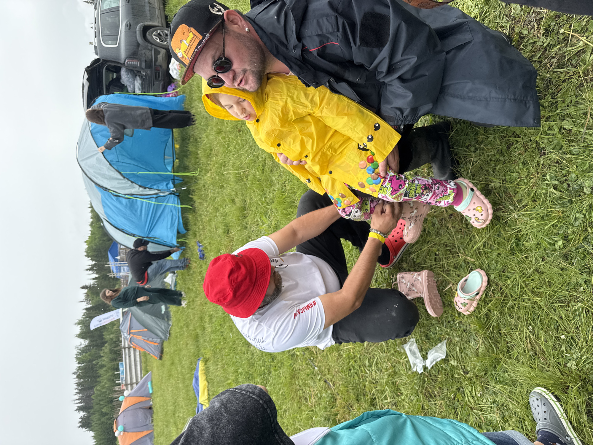 The image captures an outdoor scene at what appears to be a camping event or festival in Bol'shaya Kivara, Russia, under an overcast sky. In the foreground, a bearded man wearing a black baseball cap, sunglasses, and a black rain jacket holds a small child clad in a bright yellow rain jacket and colorful, patterned leggings. The child's yellow jacket features small black cartoon eyes and multi-colored circular shapes. Another man, wearing a red bucket hat and a white t-shirt with red text, kneels in front of them, seemingly adjusting or helping with the child's pink Crocs footwear. Multiple pairs of shoes, including other pink Crocs and brown boots, are scattered on the grassy ground nearby, along with some discarded plastic wrappers.
In the mid-ground, several camping tents of various colors (blue, grey/orange) are pitched across a green, possibly damp, grassy field. People are visible near these tents, engaged in activities like setting them up or moving around. One person in a grey jacket tends to a blue tent, while another person in a dark green top stands near a grey and orange tent. In the background, a dark-colored SUV with its trunk open is parked, suggesting arrival or packing/unpacking. The scene is bordered by a line of coniferous trees on the horizon, indicating a natural, possibly forested, setting. The overall atmosphere suggests a practical response to outdoor conditions, with rain gear being worn by some participants.
Visible text includes Cyrillic writing on the red bucket hat and a hashtag "#ЯВМБИСА" on the white t-shirt of the kneeling man. A white flag or banner in the mid-ground also has some partially visible text.