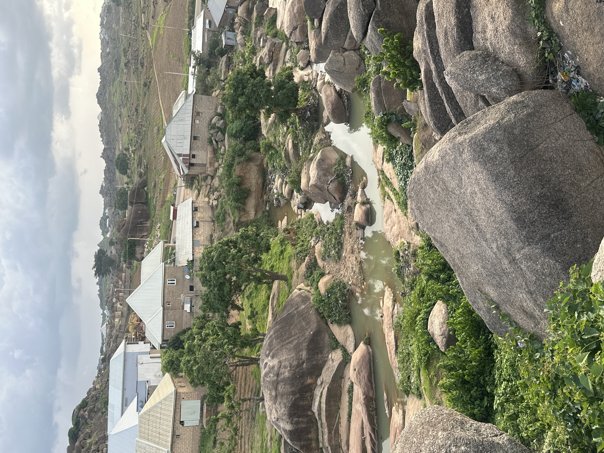 The image shows a rocky landscape in Jos, Nigeria, with scattered buildings and a small river. The buildings are primarily made of concrete blocks with corrugated metal roofs. Some of the structures appear to be houses, while others might be workshops or storage units. The river is narrow and appears to be shallow, with rocks and vegetation lining its banks. The surrounding terrain is characterized by large boulders and rolling hills covered in sparse vegetation. The sky is overcast, suggesting a cloudy or potentially rainy day. The overall impression is one of a rural or semi-rural settlement integrated into a rugged natural environment.