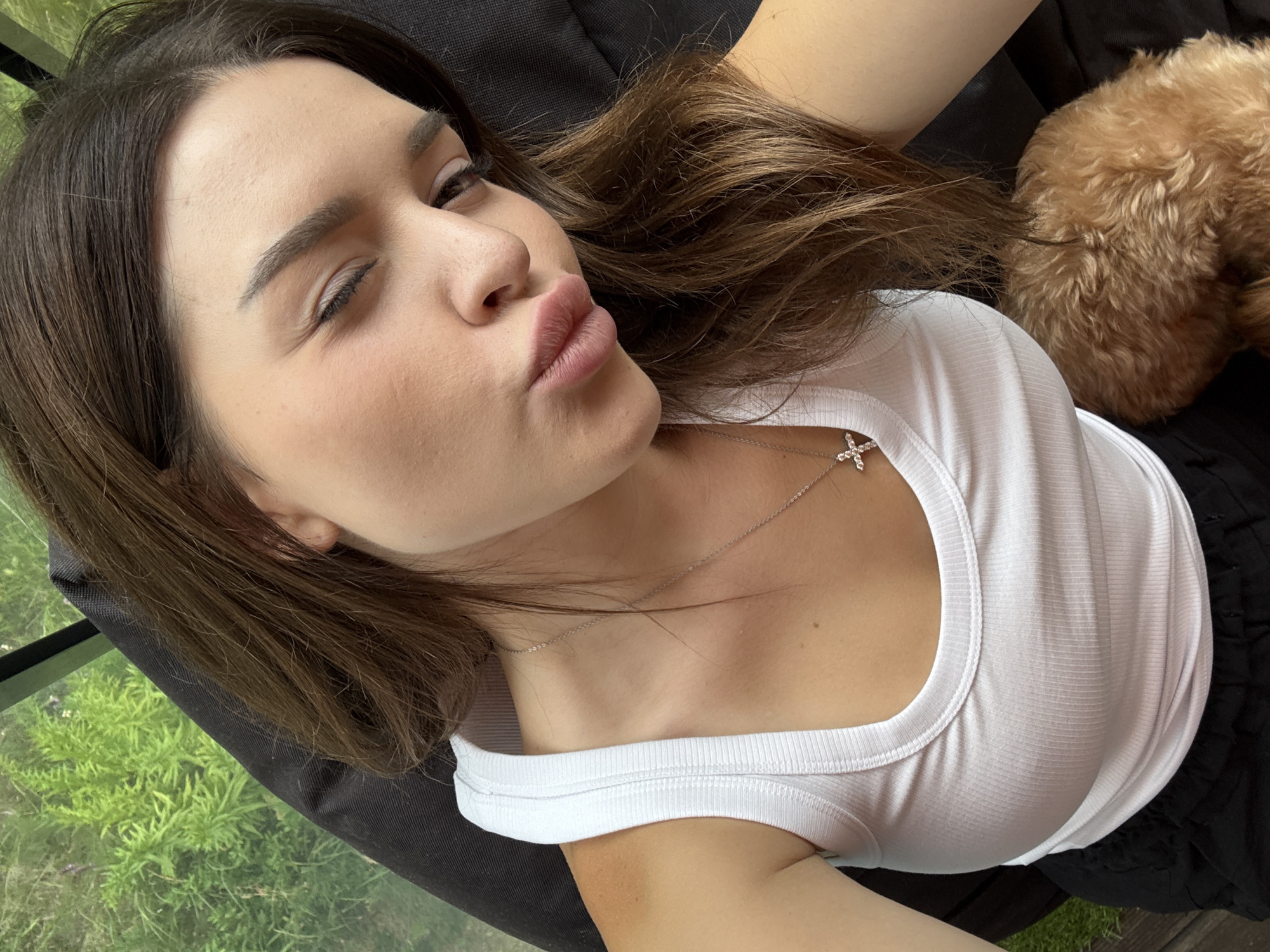 A young woman with brown hair is taking a selfie, puckering her lips and winking. She is wearing a white tank top and a silver necklace with a cross pendant. To her right, a small, fluffy dog with light brown fur is visible. The background appears to be an outdoor setting with lush green foliage, possibly a garden or park. There is no indication of Prague, Czechia in the image.