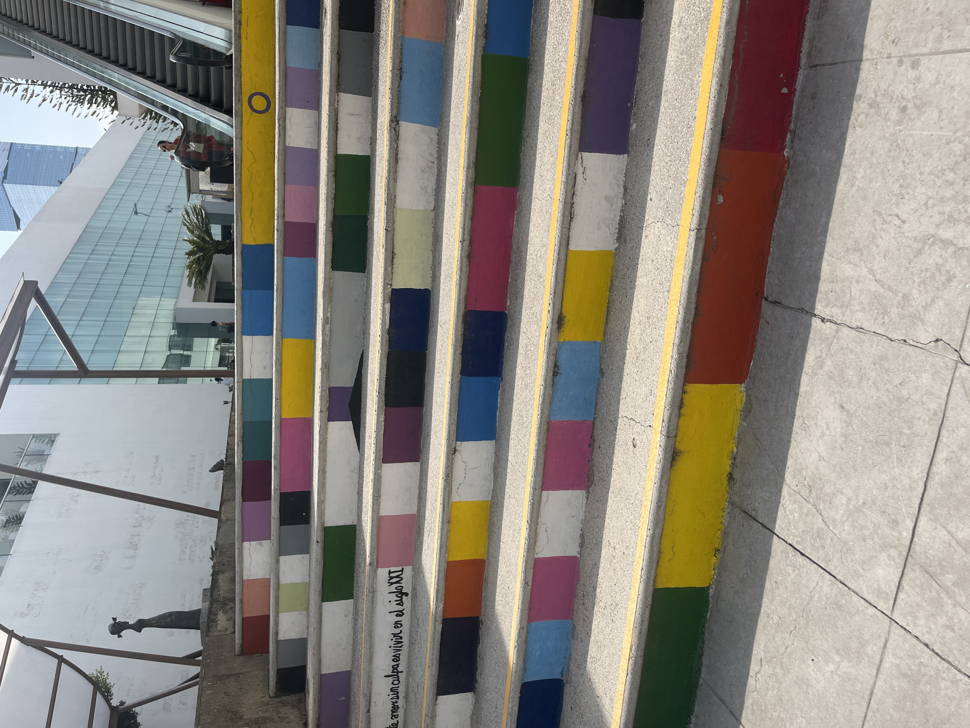 The image shows a staircase in an urban environment. The treads of the staircase are painted in a colorful, mosaic pattern. The right side of the staircase is painted with blocks of green, yellow, orange, and red. The leftmost set of treads feature blocks of yellow, blue, pink, and purple. A building with glass windows and an escalator is visible in the background. A person is seen riding the escalator. A grey wall with an ornamental sculpture and a metal railing is on the side of the staircase. Text in black is visible in Spanish on one of the stair risers. The image appears to be taken during daylight hours, casting shadows on the steps. The location is Unknown, Unknown.