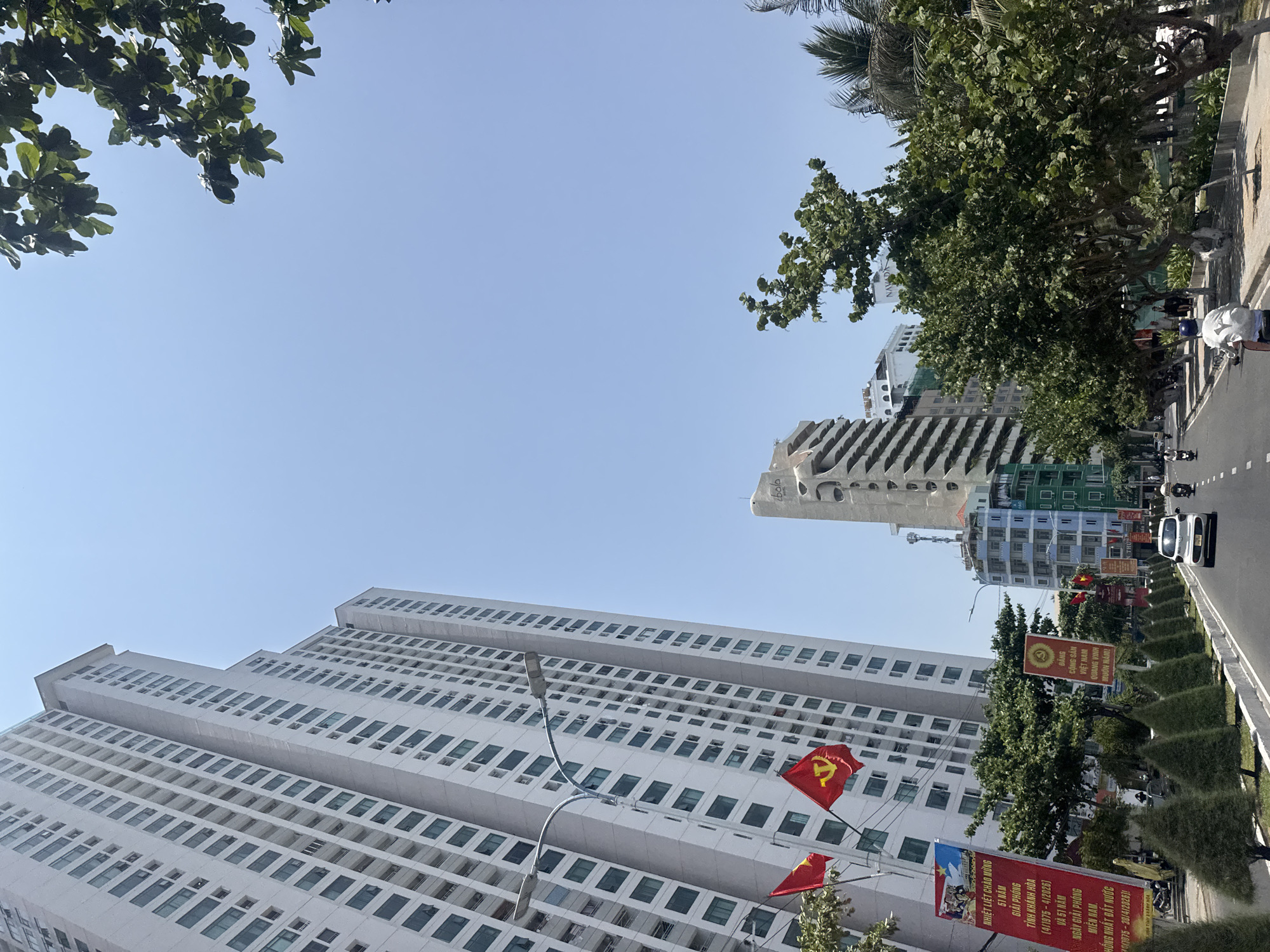 Here is a detailed description of the image:
The image shows a street scene in an urban setting, with a mix of modern architecture and greenery. In the foreground, there is a tall, white building with numerous windows, suggesting it could be an apartment complex or an office building. Two red flags with yellow stars, likely Vietnamese flags, are attached to the building.
To the right of the building is a street with a sidewalk lined with trimmed bushes. Several signs with red backgrounds and yellow text stand along the sidewalk, with the following content displayed:
- "NHIỆT LIỆT CHÀO MỪNG 51 NĂM GIẢI PHÓNG TỈNH KHÁNH HÒA (4/1975-4/2026) VÀ 51 NĂM NGÀY GIẢI PHÓNG MIỀN NAM. HỒNG NHẤT ĐẤT NƯỚC"
- "BANG CỘNG SẢN VIỆT NAM QUANG VINH MUÔN NĂM"
A few vehicles are visible on the road, including what appears to be a white car. People on motorcycles can be seen in the distance, suggesting active street traffic.
In the background, there are more buildings, including a uniquely designed, light-colored structure with curving lines. Trees are present, both along the street and framing the top of the image, adding a touch of nature to the urban environment. The sky is clear and blue.
