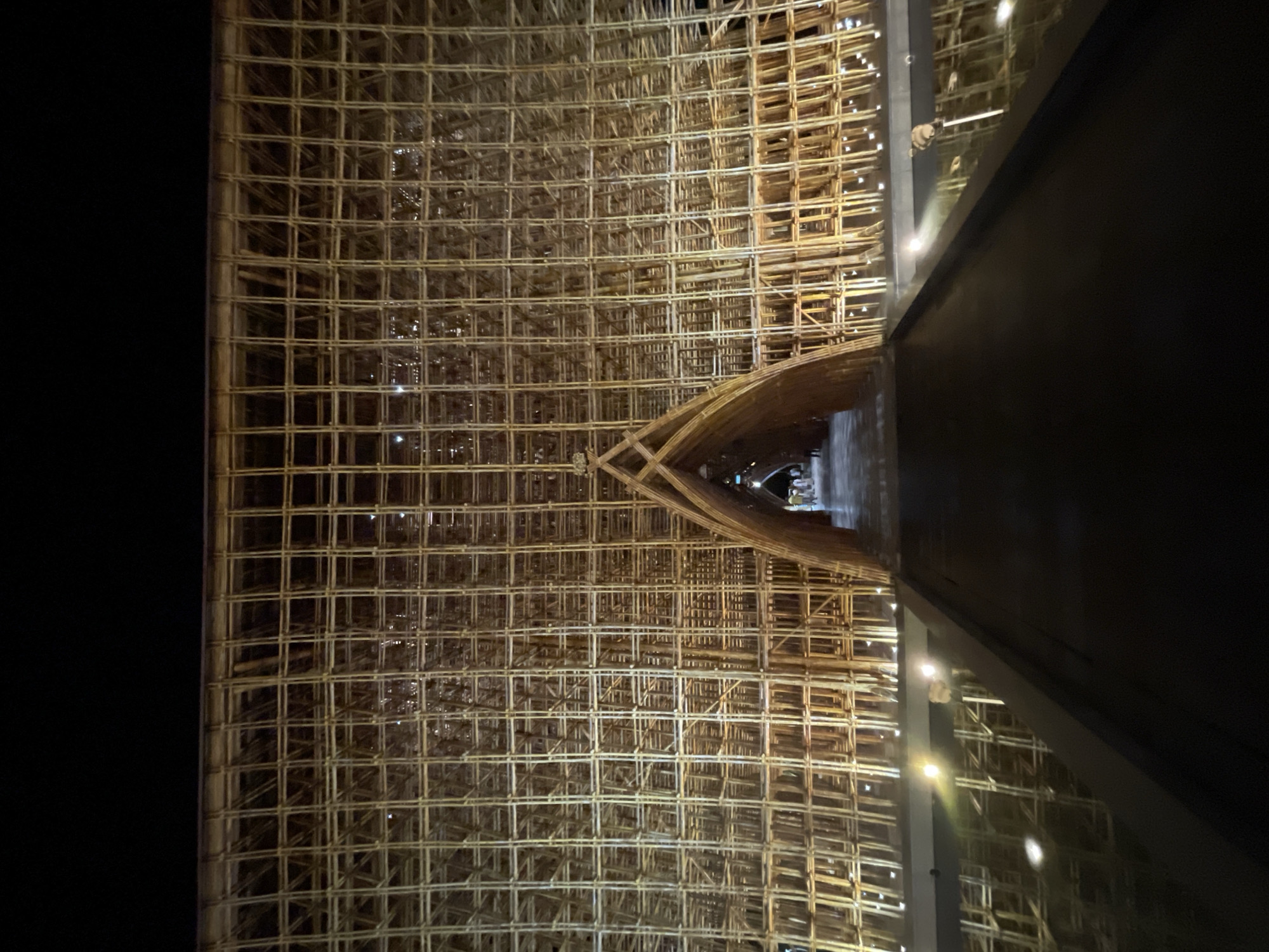 The image displays a grand architectural structure at night, dominated by an immense, intricately woven grid-like wall, likely constructed from bamboo or light wood. The structure fills most of the frame, showcasing countless small, square and rectangular openings forming a complex, textured pattern.
In the middle-right section of the grid wall, a prominent archway or tunnel entrance is visible, also made from the same dense framework, leading into a dimly lit interior space. Tiny figures of people can be seen within this passage, indicating the significant scale of the structure.
To the right, a large, dark, and highly reflective surface captures the illumination from within and around the grid, showing reflections of the patterned framework and numerous bright, artificial light sources. These lights cast a warm glow on the wooden structure, enhancing its geometric details and creating a dramatic contrast against the surrounding darkness. The scene is clearly set at night, with artificial lighting being the primary source of illumination. The elaborate and organic construction suggests a unique architectural installation or landmark, possibly in Phú Quốc, Vietnam. No visible text is present in the image.