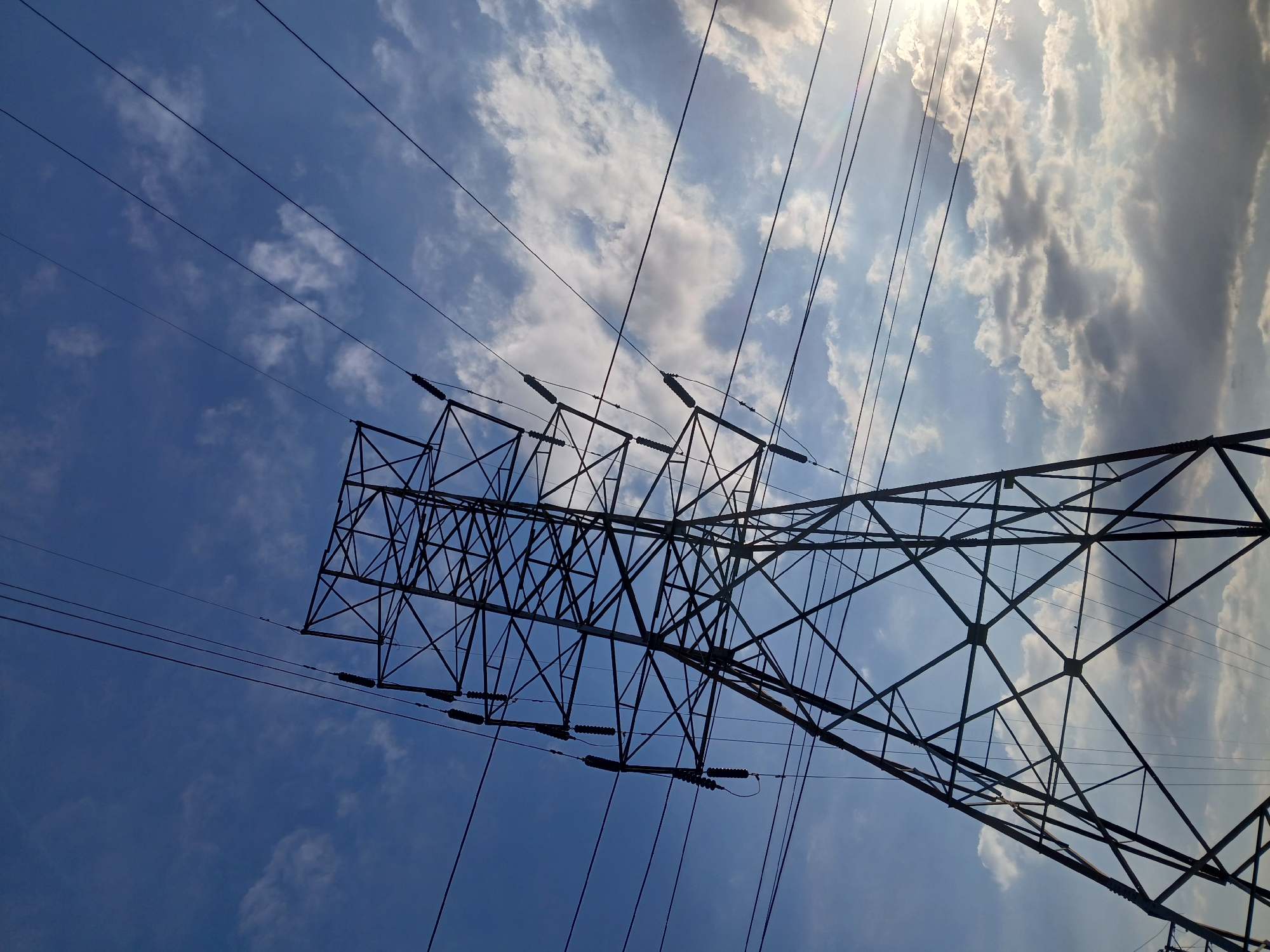The image shows a low-angle view of an electricity pylon against a sky with scattered clouds. The pylon is made of metal and has a lattice structure. Electrical wires extend from the pylon. The sky is blue with some white clouds, and the sun is shining brightly. The location is Ciudad Guayana, Venezuela.