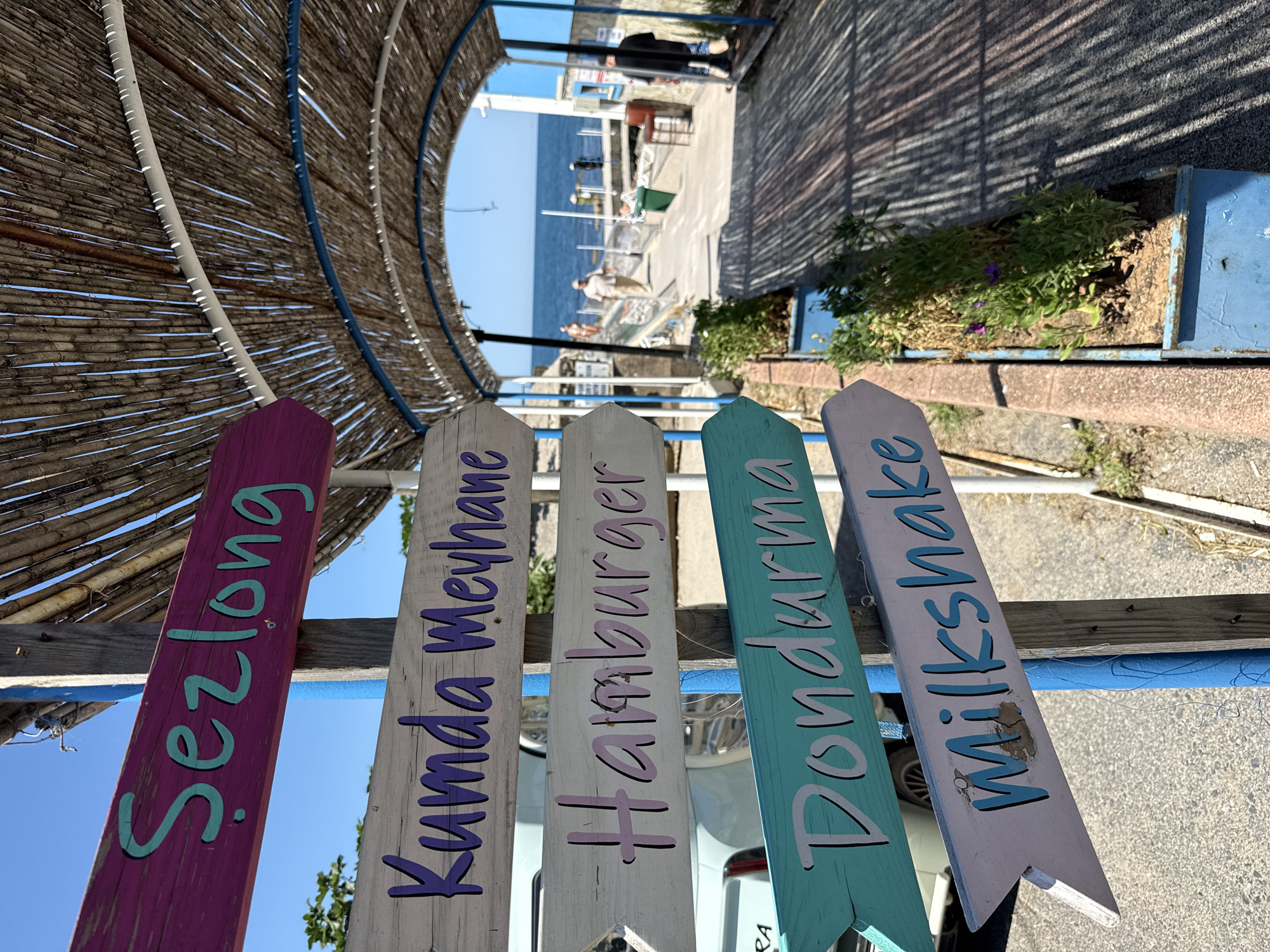 The image shows a directional signpost with several wooden arrow-shaped signs in the city of Şile, Turkey. The signs are painted in different colors and display various food and beverage options in Turkish and English.
From top to bottom, the signs read:
1. "Sezlong" (Turkish for sun lounger), painted purple with turquoise lettering.
2. "Kumda Meyhane" (Turkish for "Tavern on the Sand"), painted white with purple lettering.
3. "Hamburger", painted white with pink lettering.
4. "Dondurma" (Turkish for ice cream), painted turquoise with white lettering.
5. "Milkshake", painted white with turquoise lettering.
The signpost is located outdoors, near a pathway that leads to the sea. In the background, people can be seen walking towards the water, and the sea and a clear blue sky are visible. The pathway is partially covered by a thatched roof structure, creating a shaded area. The overall scene suggests a casual, beachside dining or recreational area.