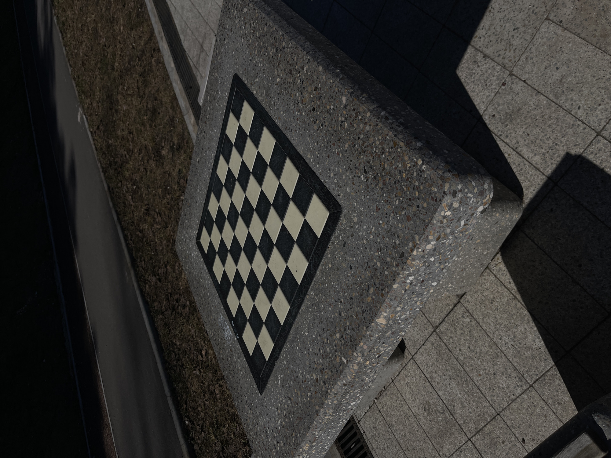 A high-angle view shows a gray, stone chessboard embedded in a matching stone bench located in Chaikovsky, Russia. The chessboard displays a standard black and white square pattern. The bench is situated on a tiled surface with noticeable shadows. A strip of dry, brown ground, possibly a garden bed, is visible adjacent to the bench. A dark, smooth surface, likely a road or path, is on the opposite side of the garden bed. The lighting is bright, emphasizing the textures of the stone and the contrasts between the various surfaces. No people are present in the frame.