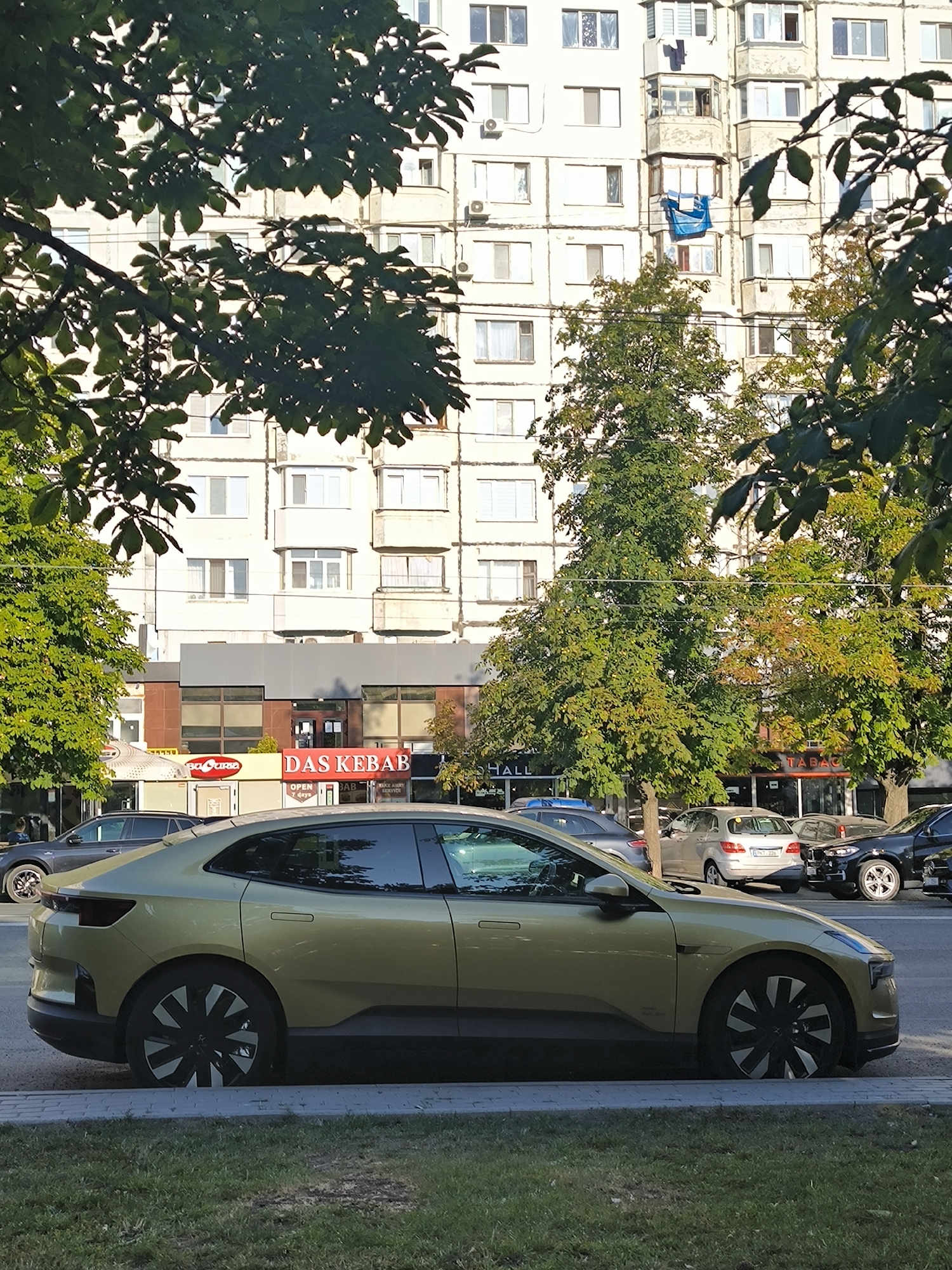 The image shows a light gold electric car parked on the side of a street in Chișinău, Moldova. The car is positioned in front of a modern building with several shops on the ground floor, one of which is labeled "DAS KEBAB". Behind the shops, a multi-story apartment building with light-colored facades is visible. Trees with green foliage partially obscure the buildings. The car is the primary subject, with its side profile clearly displayed. The sky is a clear, light blue. The scene appears to be a typical urban environment on a sunny day.