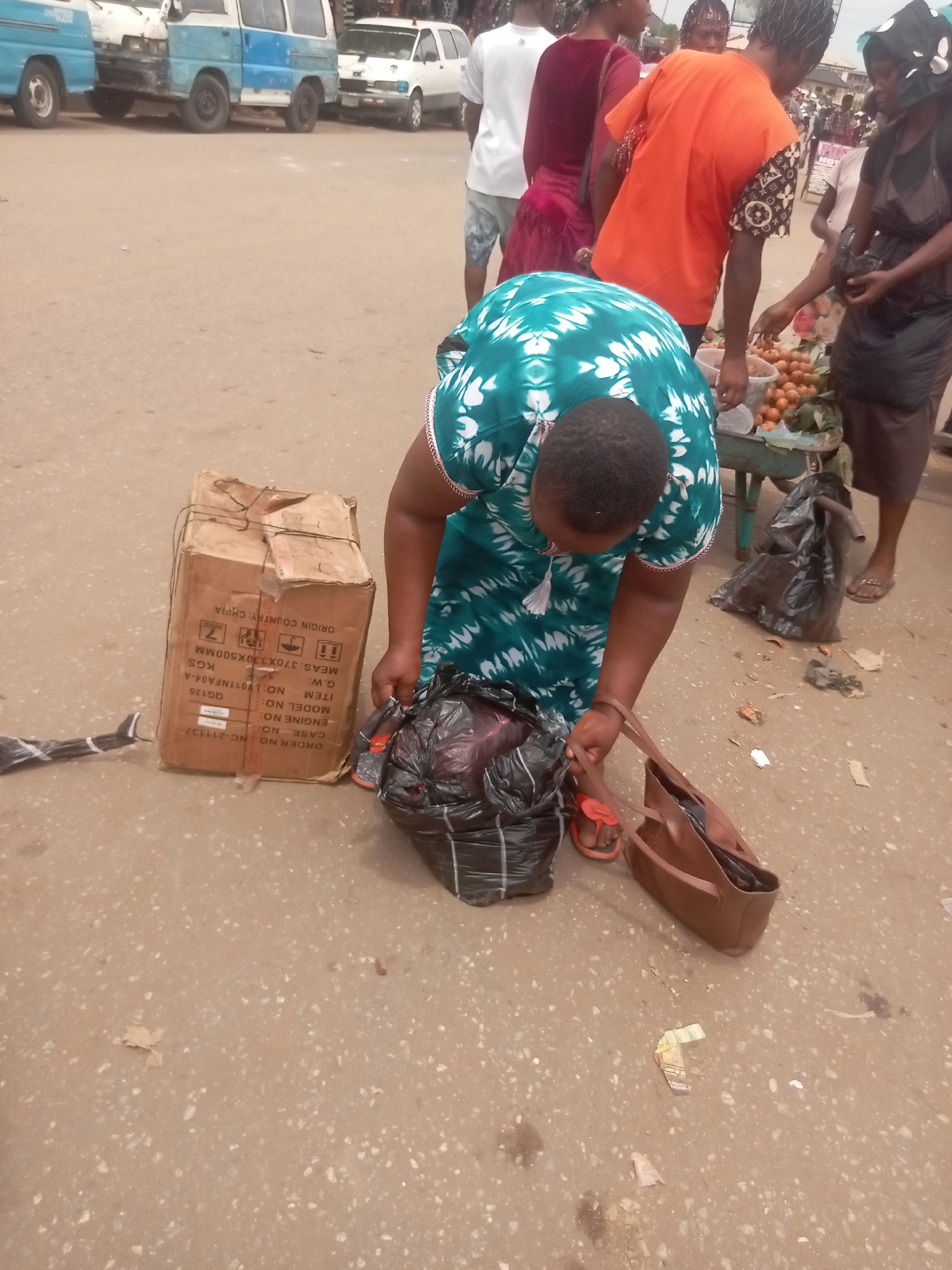 A woman in a green patterned dress bends to place a plastic bag on the ground. Next to the bag is a brown leather purse and a cardboard box, which has shipping labels printed on it stating "ORIGIN COUNTRY: CHINA". A few other people and street vendors surround her. Several minibuses are parked along the street. The scene appears to be an outdoor market location.