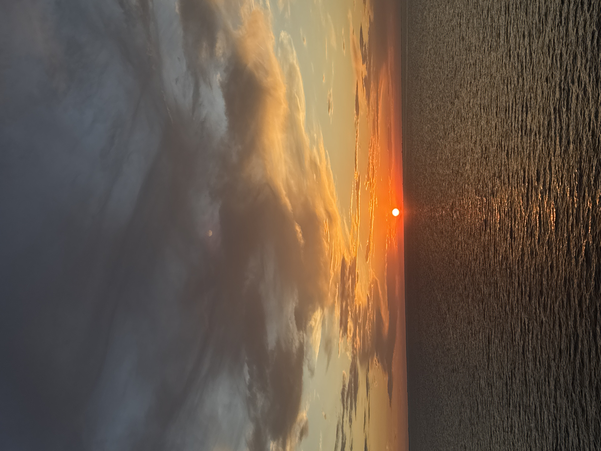 An extreme wide shot captures a sunset over the ocean. The sun, a bright orange orb, is low on the horizon, casting a warm glow on the sky and water. Wispy clouds, painted in shades of orange, pink, and gray, stretch across the sky. The ocean is a deep blue, with gentle waves reflecting the sunset's colors. The location is the sea, with no identifiable landmarks in the city of Unknown, Italy.