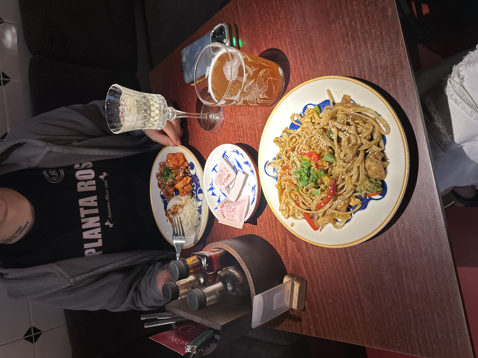 The image shows a table at a restaurant, likely in Moscow, Russia, with two plates of food and two drinks. The food consists of noodles with vegetables and meat, and a dish of chicken with rice. There are also two bottles of sauces in a wooden holder. The drinks include a glass of white wine and a glass of beer. A person, wearing a grey hoodie and a black t-shirt with the logo "PLANTA ROSA", is partially visible in the background, sitting at the table. The table is made of dark wood, and the background is a dark, possibly upholstered booth seat. The overall setting suggests a casual dining experience.