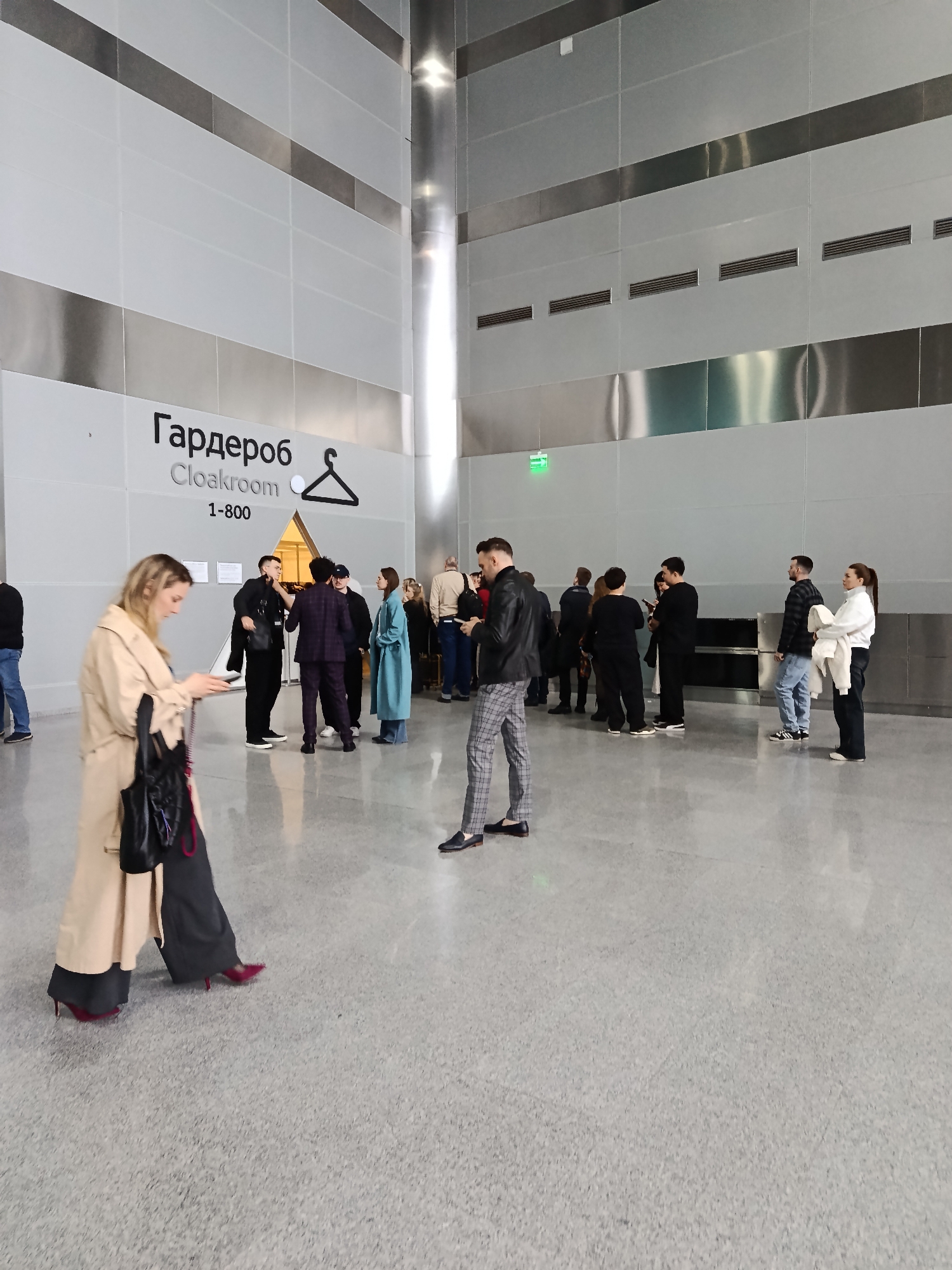 The image depicts the interior of a modern building with a line of people queuing near a "Гардероб Cloakroom" sign. The sign also includes an icon of a coat hanger and the number 1-800. The interior has gray tiled floors and walls, with a vertical silver accent running from floor to ceiling. Several people are standing in line, predominantly dressed in dark clothing. A woman in the foreground is wearing a light beige trench coat and dark trousers with red heels, looking at her mobile phone. The location is likely a public space in the city of Khimki, Russia, given the Cyrillic signage.