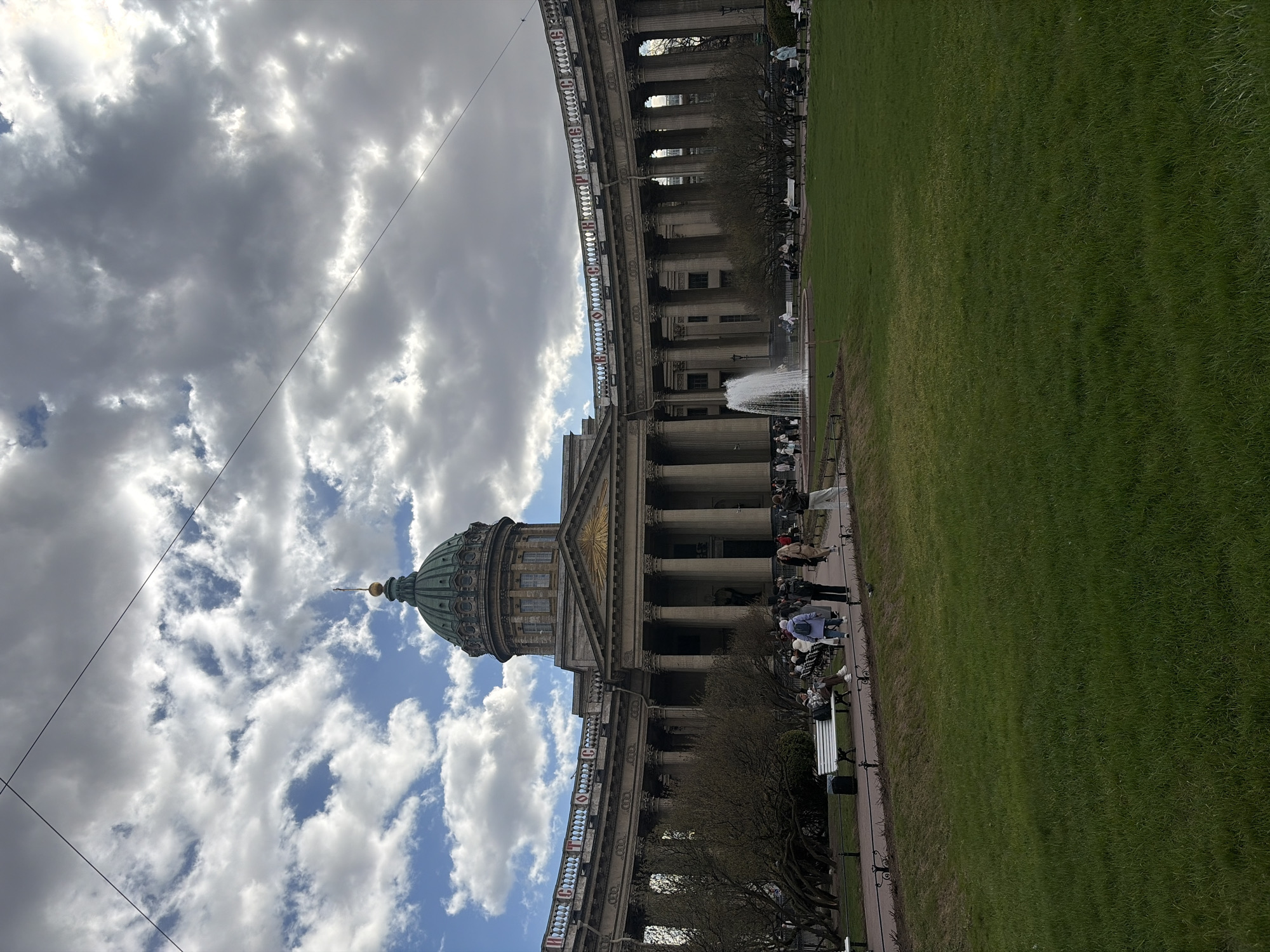 The image depicts the Kazan Cathedral in Saint Petersburg, Russia. The cathedral is captured from a low angle, showcasing its dome and colonnades. The sky above is filled with clouds, ranging from dark and ominous to bright and partly sunny. A power line stretches across the sky in the foreground. People are seen around the cathedral, some sitting on benches and others walking. A large green lawn extends to the right of the cathedral. A fountain is visible in front of the building.