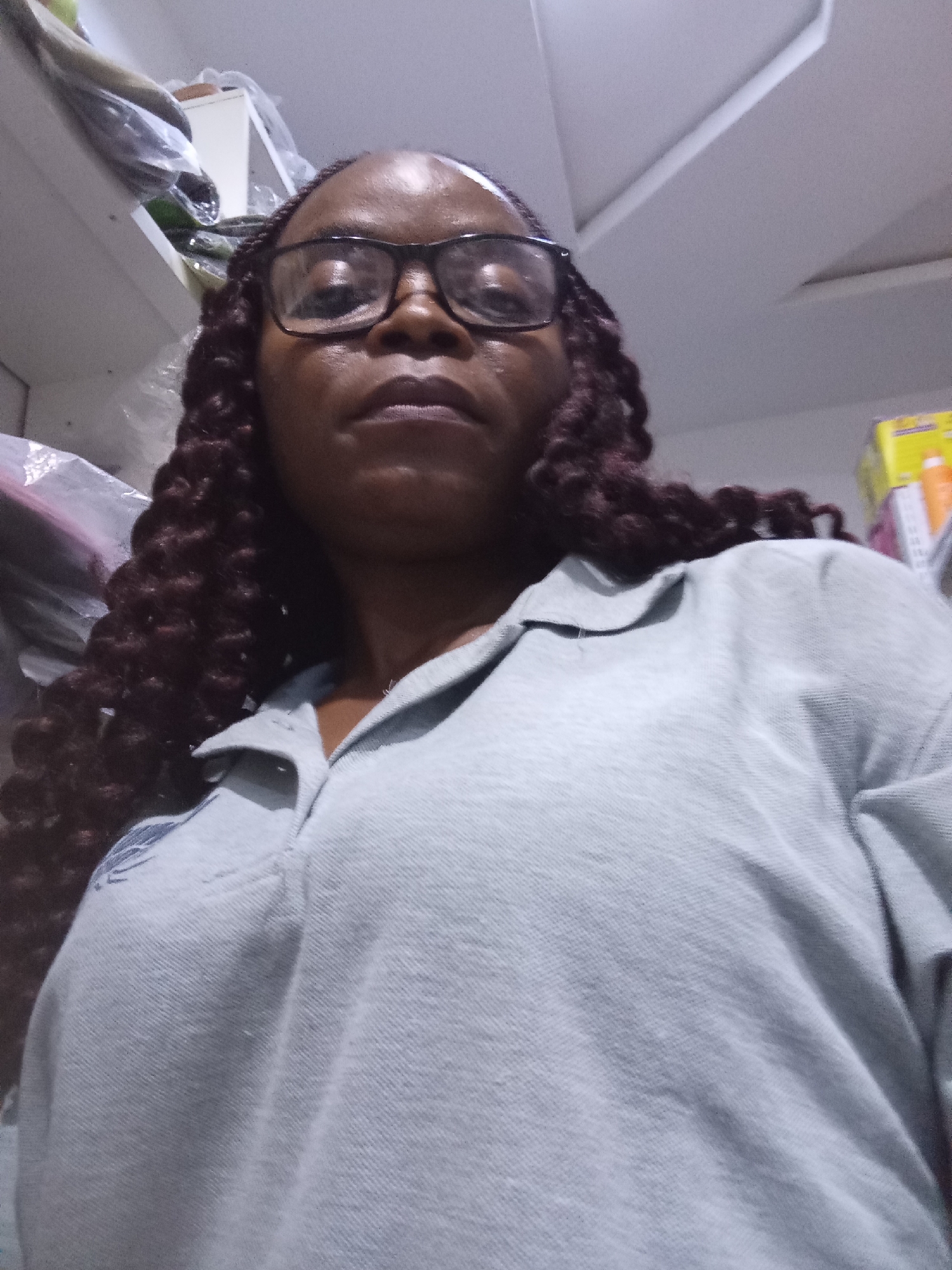 A woman with dark skin and dark curly braided hair, wearing glasses and a light grey polo shirt, is pictured in a selfie. The camera angle is from below, looking up at her face, which is slightly tilted. Her expression is neutral. In the background, shelves are visible, some with items on them, and a white ceiling with angled panels. The scene appears to be indoors, possibly in a room or storage area, and does not contain identifiable landmarks of Howayi, Nigeria.