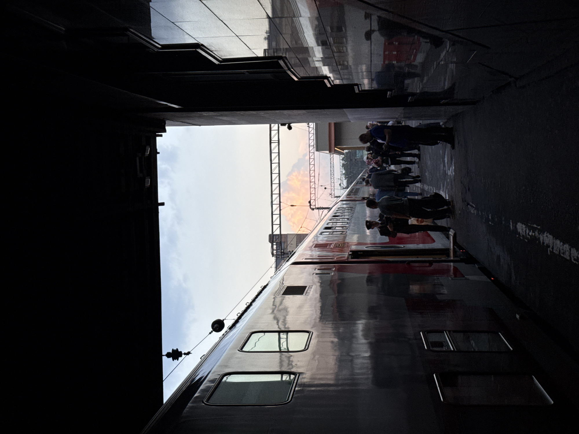 The image shows the side of a train at a station. People are standing alongside the train. The photo is taken from a dark, possibly enclosed, location, looking out towards the train and platform. The sky is visible in the distance. There is no definitive evidence to confirm the location as Ukromne, Ukraine.
