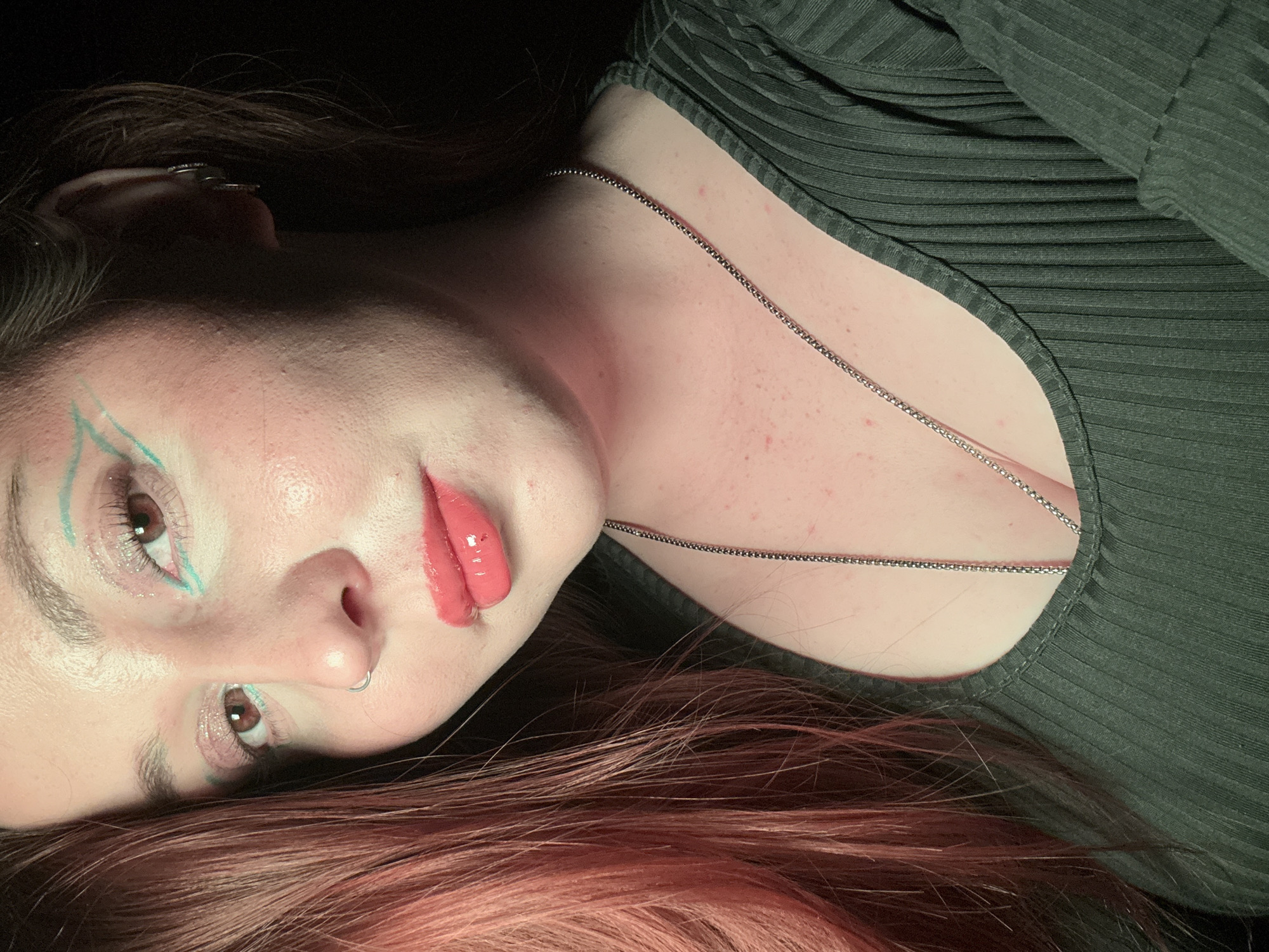 A close-up, angled portrait features a fair-skinned individual with light brown eyes and auburn-tinted hair resting against a dark backdrop. The individual is adorned with teal eyeliner extending from the inner corner of each eye, and a glossy coral lip color. A silver nose ring is visible. The individual wears a ribbed, dark green top with a scoop neckline, and a thin silver chain rests on the chest. The surrounding environment is dark and undefined. The location of the image within the city of Izhevsk, Russia cannot be determined.