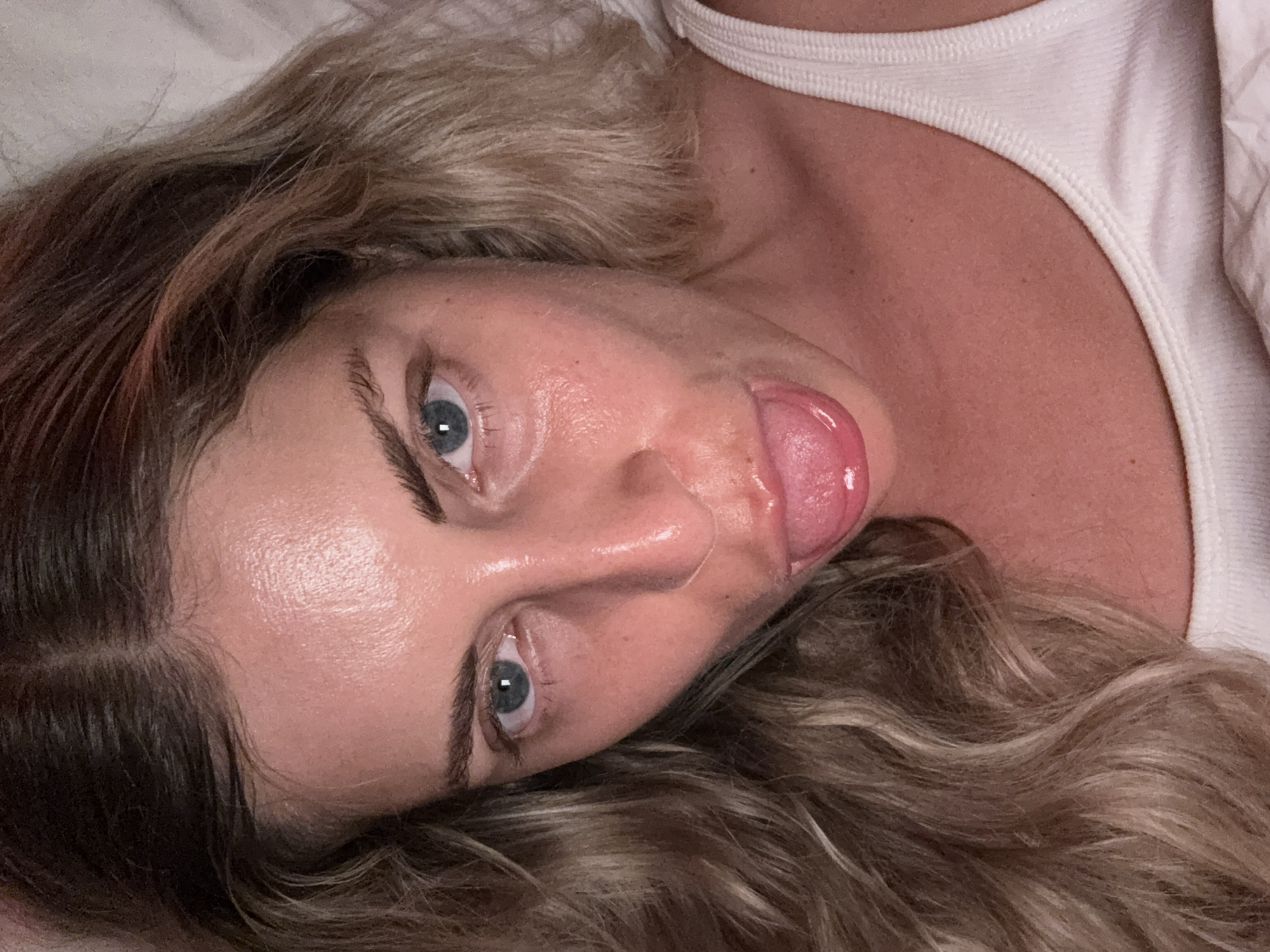 A close-up selfie of a woman with blonde wavy hair, lying down. She is looking directly at the camera with her eyes open and tongue sticking out. Her skin is slightly shiny, and she is wearing a white tank top. The background is blurry, suggesting an indoor setting. There is no indication of the location being Paris, France.