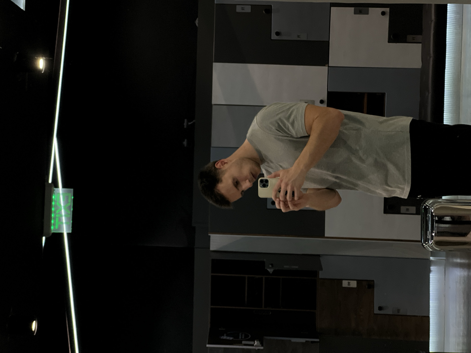 A person with short dark hair stands in front of a mirror, holding a cell phone in their hands. The person wears a gray t-shirt and black pants. The reflection in the mirror shows a backdrop of storage lockers that are shades of gray and white. Adjacent to the mirror's reflection is a section of wall, mostly black with a green "EXIT" sign affixed to it. There is no information in the image to verify the location in Burgas, Bulgaria.
