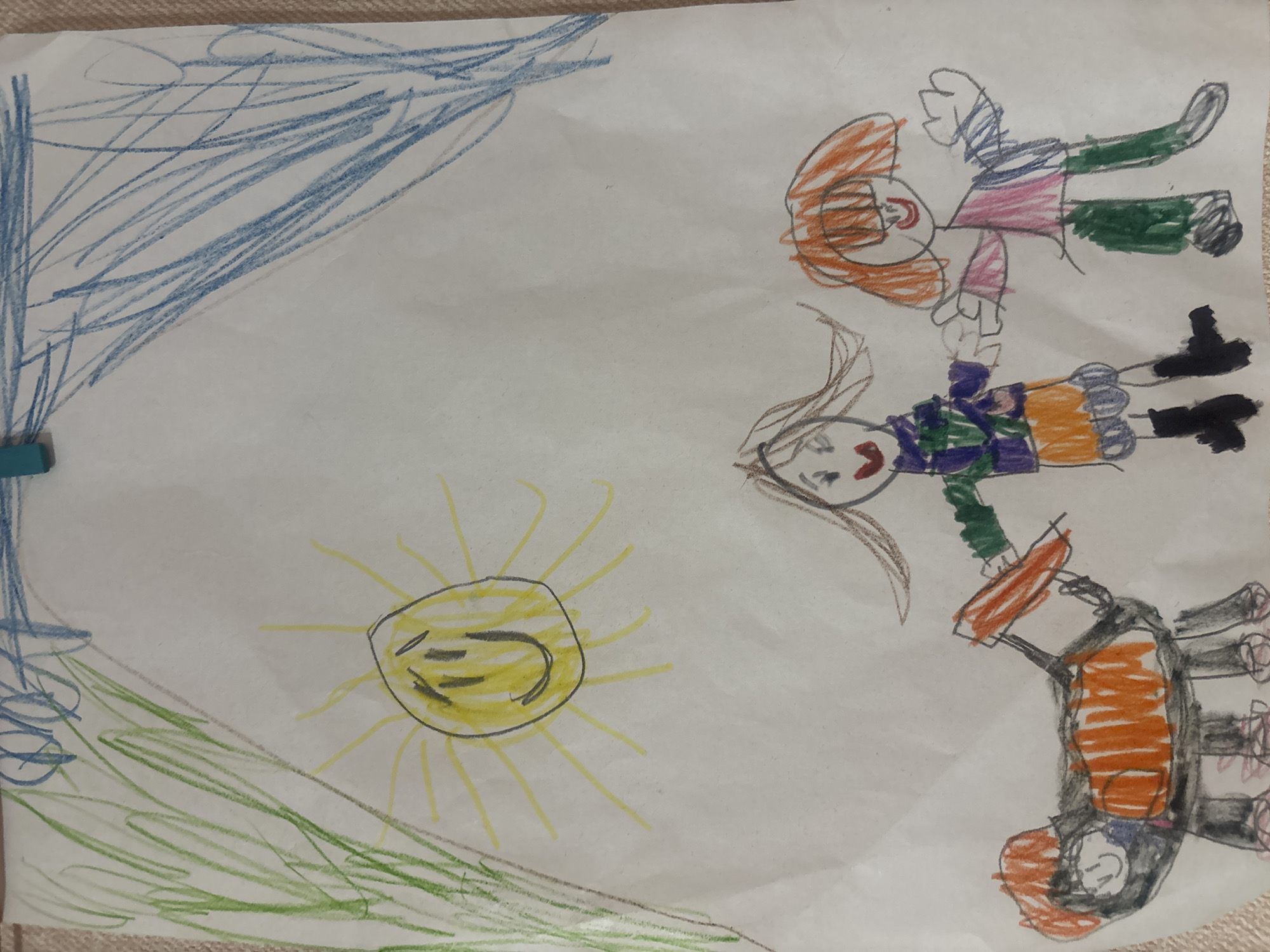 This is a child's drawing on white paper. The drawing depicts a sunny outdoor scene with two figures and an animal. In the upper portion of the drawing, a yellow sun with a smiley face and radiating lines is visible. To the left of the sun, green scribbles represent grass or trees, and to the upper right, blue scribbles suggest a sky or more foliage.
In the lower part of the drawing, two stick figures, presumably children, are interacting with a four-legged animal. The figure on the left has brown hair and is wearing a purple and green jacket over an orange skirt, with black boots. The figure on the right has orange hair and is wearing a pink shirt and green pants. They appear to be holding hands or interacting with each other.
The animal is black with orange markings and has four legs. One of the figures is holding an orange object that resembles a tool, possibly grooming the animal. The overall style is characteristic of early childhood art. The image is pinned to a surface with a blue clip at the top. The location of Yanino-1, Russia, is not depicted or suggested by any elements within the drawing itself; this information is external to the visual content of the image.