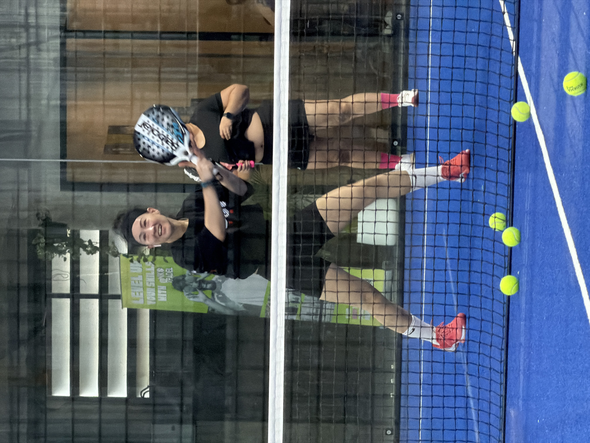 Two women are playing padel on an indoor blue court in Ban Buak Khrok North, Thailand. The woman in the foreground, on the left, is wearing a black headband, black t-shirt, and black shorts. She has on white socks and red sneakers. She is smiling and holding a padel racket in her right hand, extended towards the other player. The woman in the background, on the right, is wearing a black t-shirt and black shorts. She has on pink socks and white sneakers. She is holding a padel racket over her head. The court has a white net across the middle. Several yellow padel balls are visible on the court. In the background, there is a green banner with text that says "LEVEL UP YOUR SKILLS WITH PADEL". There is also a window with horizontal blinds, and plants hanging from the ceiling.
