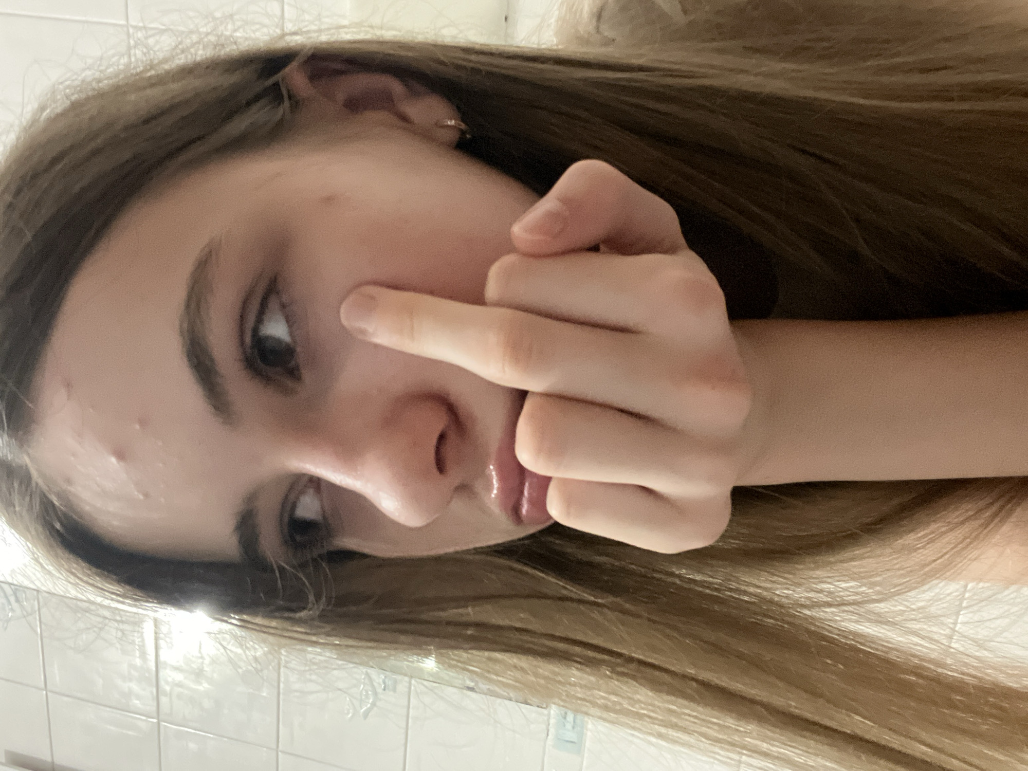The image shows a close-up, profile view of a young Caucasian female with long, light brown hair. She is resting her chin on her hand, with her index finger touching her cheekbone. Her eyes are looking off to the left, and her lips are slightly parted and appear to be glossed. There are visible blemishes on her forehead. She is wearing a small hoop earring in her right ear. The background appears to be a tiled wall, suggesting an indoor setting. No specific location in Dnipro, Ukraine can be identified from the visual information provided. The lighting suggests it may be artificial.