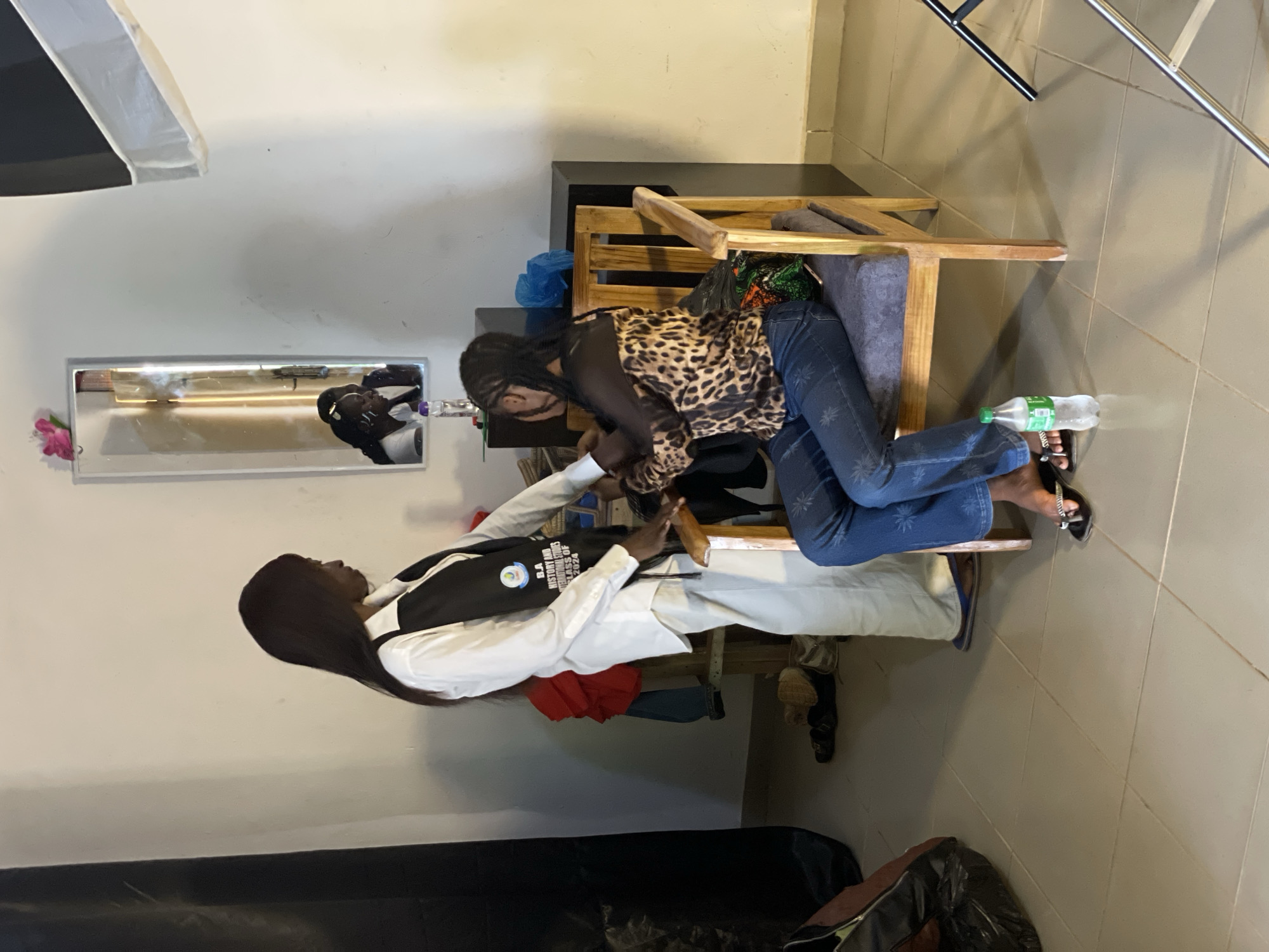 Two individuals are present in the scene, with one person seated on a wooden chair and the other standing. The seated person, wearing a leopard-print top and blue jeans, is leaning forward. The standing person is dressed in a white shirt and a black vest, with text visible on the vest. Both individuals appear to be interacting, with their hands engaged in an activity that is not fully discernible.
The setting seems to be an indoor space with tiled flooring. A mirror is mounted on the wall, reflecting a portion of the room and one of the subjects. Various objects are scattered around, including a bottle of water on the floor, a pink flower near the mirror, and some bags. A large black object, possibly a piece of equipment or furniture, occupies the lower part of the frame.
The lighting suggests it might be daytime, with artificial lights also contributing to the illumination. The overall atmosphere appears casual, and there are no strong indicators of specific emotions. The text on the vest reads "B.A HISTORY AND INTERNATIONAL STUDIES CLASS OF 2024". The location context provided is Durumi, Nigeria.