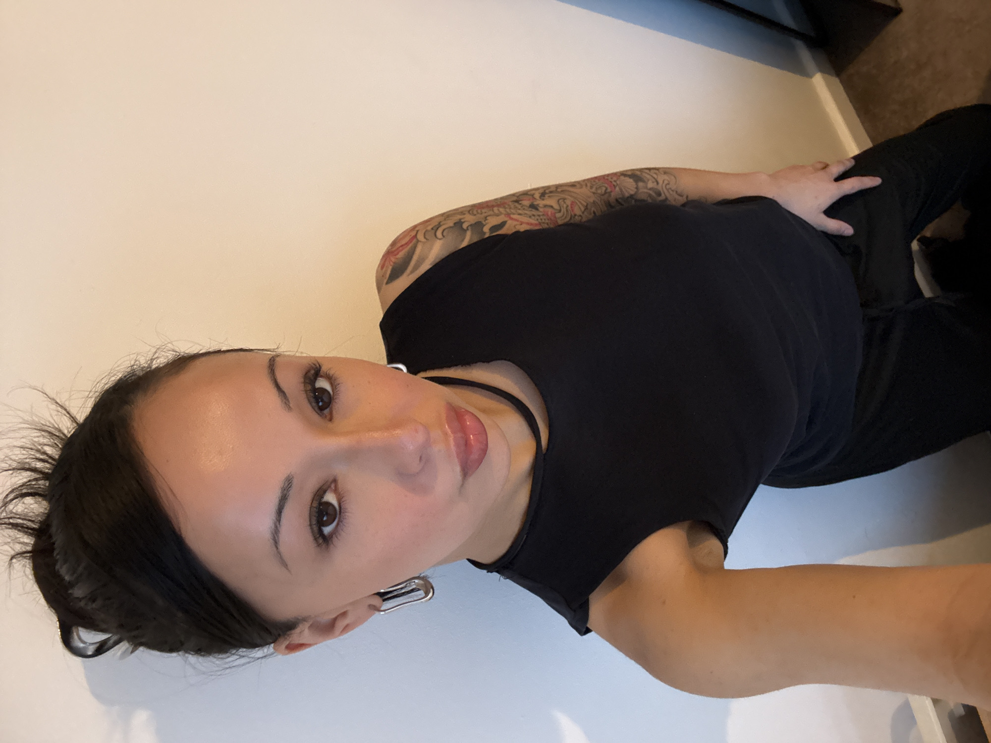 A woman is centered in a waist-up, close-up shot. Her dark hair is pulled back from her face, and she has dark eyes and full lips. She is wearing a black top and a black choker necklace. A large, silver earring hangs from her left ear. Her left arm, which is extended, is visible at the bottom right of the frame, while a tattoo is visible on her upper left arm. She is lying on the side, angled toward the camera. The background consists of a plain, light-colored wall, and what appears to be the corner of a dark piece of furniture.
There is no indication of the location being in the city of London, United Kingdom.