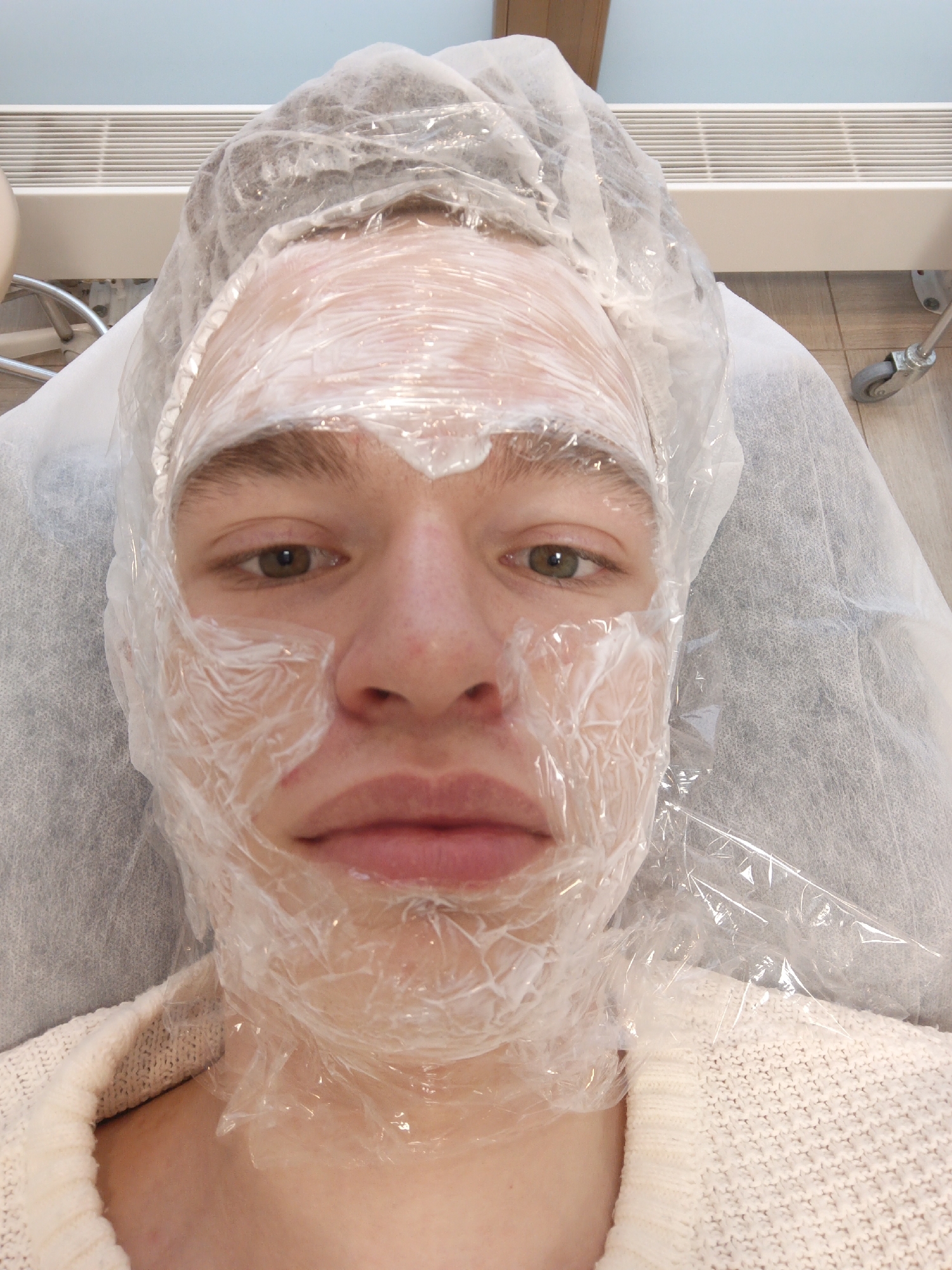 A young, light-skinned person lies on a padded examination table covered with a white disposable sheet. They are wearing a white sweater and have a clear plastic wrap applied over a white substance on their face, and a clear plastic hairnet. The person's face is visible, with brown hair, brown eyes and pink lips. In the background is a white wall-mounted heater with horizontal slats and a wood-grain floor. There is also a portion of a wheeled examination table or chair. There is no information to support the location being in the city of Moscow, Russia.