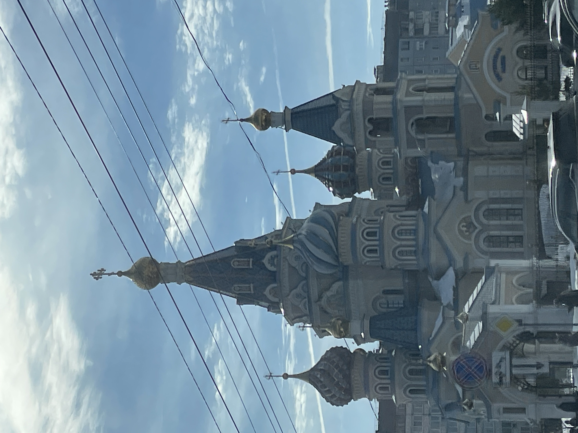 The image displays the Cathedral of the Holy Archangel Michael in Izhevsk, Russia, captured under a partly cloudy blue sky. The cathedral features a multi-domed structure with a dominant central spire culminating in a golden onion dome and an Orthodox cross. The architectural style incorporates light beige and pale blue elements, with darker blue roofing on some sections and several smaller, intricately patterned blue and white domes. Numerous golden onion domes, each topped with an Orthodox cross, adorn the various towers. Patches of snow are visible on the cathedral's ledges and lower roofs, indicating a winter environment. Multiple dark electrical or tram lines span diagonally across the upper portion of the frame, partially obscuring the sky. In the foreground, portions of dark-colored vehicles are present on a snow-dusted ground. Indistinct human figures are visible at ground level near the cathedral's entrance, which features a circular blue traffic sign with a red diagonal cross and a yellow diamond-shaped traffic sign. Multi-story urban residential or commercial buildings are discernible in the background, framing the cathedral. The scene is illuminated by bright daylight.
