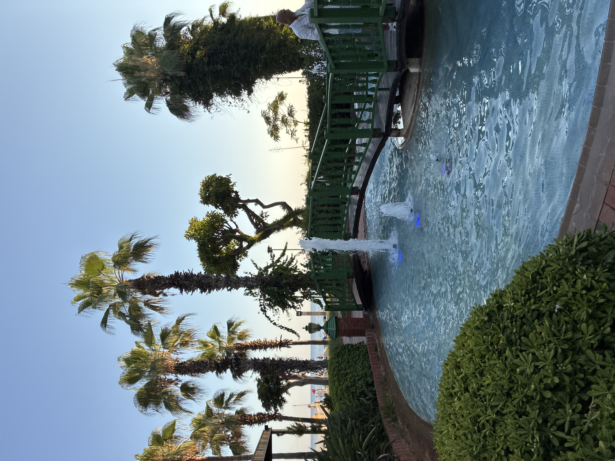 The image depicts a serene waterscape with a green arched bridge spanning a channel of clear blue water. Fountains are active in the water, creating playful sprays. Lush green bushes line the edge of the channel, and palm trees are abundant in the background, suggesting a tropical or subtropical climate.
In the middle ground, a man in a white shirt and light-colored pants stands on the bridge, looking towards the right side of the frame. The bridge has dark wooden planks and bright green railings. Behind the bridge, a beach with umbrellas and people can be seen, with the sea and the horizon visible beyond.
The scene is illuminated by soft, natural light, likely from the late afternoon or early morning sun. The overall atmosphere is peaceful and inviting.
This scene is likely located in Alanya, Turkey, given the prevalence of palm trees, the proximity to the sea, and the architectural style which is common in resort areas of the region. The presence of a well-maintained water feature and bridge suggests it could be part of a hotel, resort, or public park area within Alanya.