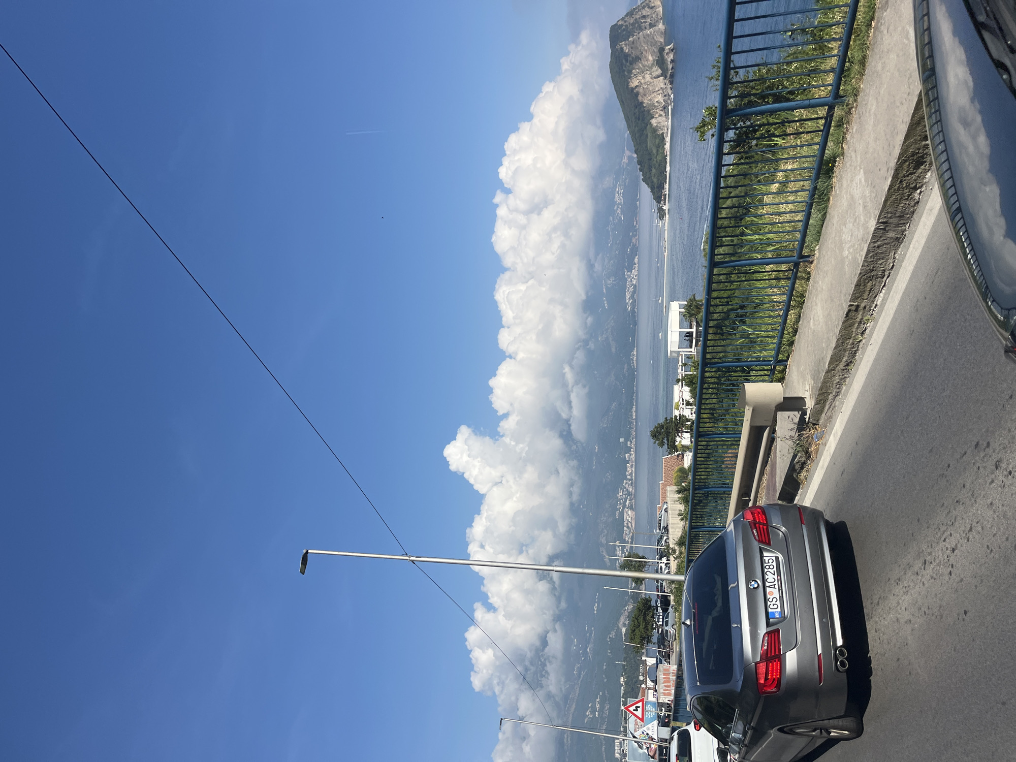 This image captures a vibrant coastal landscape viewed from a roadside. The scene is dominated by a clear blue sky, a vast body of water, and impressive mountains under a bright sun.
In the immediate foreground on the right, a grey BMW sedan (likely a 5 Series) is visible from behind. Its license plate, "GS AC285," features an "HR" sticker, indicating it's registered in Croatia. The car is on an asphalt road, and a blue metal barrier runs alongside it, separating the road from the precipitous drop to the water below. Further down the road on the left, a red triangular warning sign indicating a winding road is partially visible.
Beyond the barrier, a large bay or sea stretches out, with numerous boats and coastal towns lining the distant shores. Towering, rugged mountains rise from the water on the left, partially enveloped by large, fluffy white cumulus clouds that dramatically fill the mid-ground. The sky above is a brilliant blue, with a faint contrail of an aircraft in the upper left. Street light poles and overhead power lines traverse the upper portion of the frame.
The image suggests a clear and sunny day, likely late morning or early afternoon, given the strong illumination. The overall setting is that of a scenic drive along a coastal road. Although the license plate suggests Croatia, the provided context specifies the location as Kubasi, Montenegro.