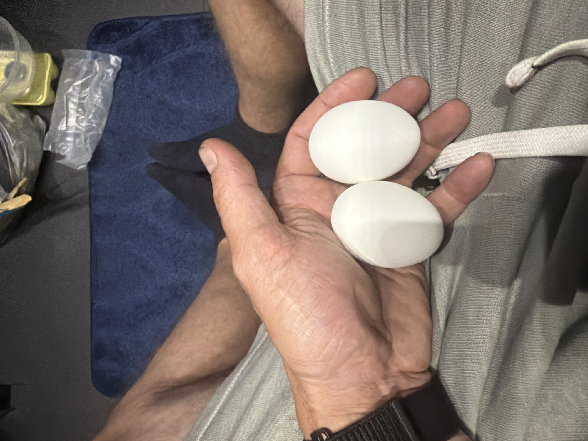 The image shows a person holding two white, oval objects in their right hand. The objects appear to be eggs. The person is wearing grey sweatpants and a black watch on their left wrist. In the background, a blue mat and a person's feet wearing black socks are visible. There are also some plastic bags and a yellow bottle in the background. The scene appears to be indoors, possibly in a living room or bedroom. The location in the city of New Britain, United States of America is not specified.