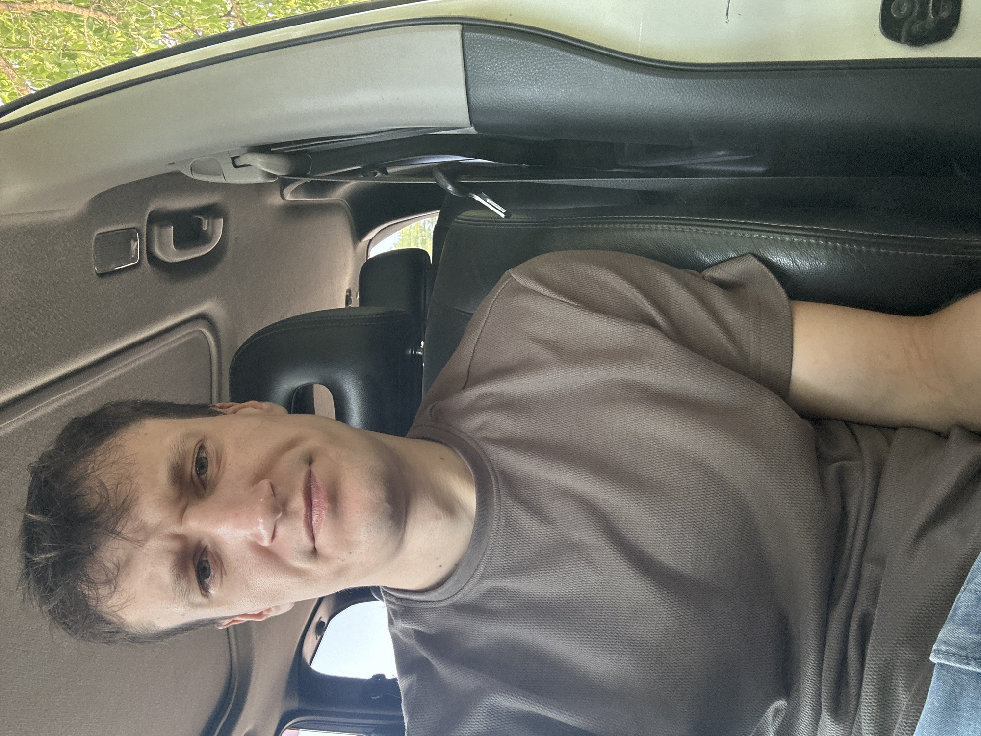 The image shows a man sitting in the passenger seat of a vehicle. He is wearing a brown t-shirt and blue jeans. The interior of the vehicle is visible, including the headrest of the driver's seat, the sunroof, and the door panel. Outside the vehicle, trees and foliage can be seen through the windows, indicating an outdoor setting, likely a rural or suburban area. The man has a neutral expression on his face. The image does not provide any specific information about the location within the city of Makiv, Ukraine, or any discernible actions or interactions.